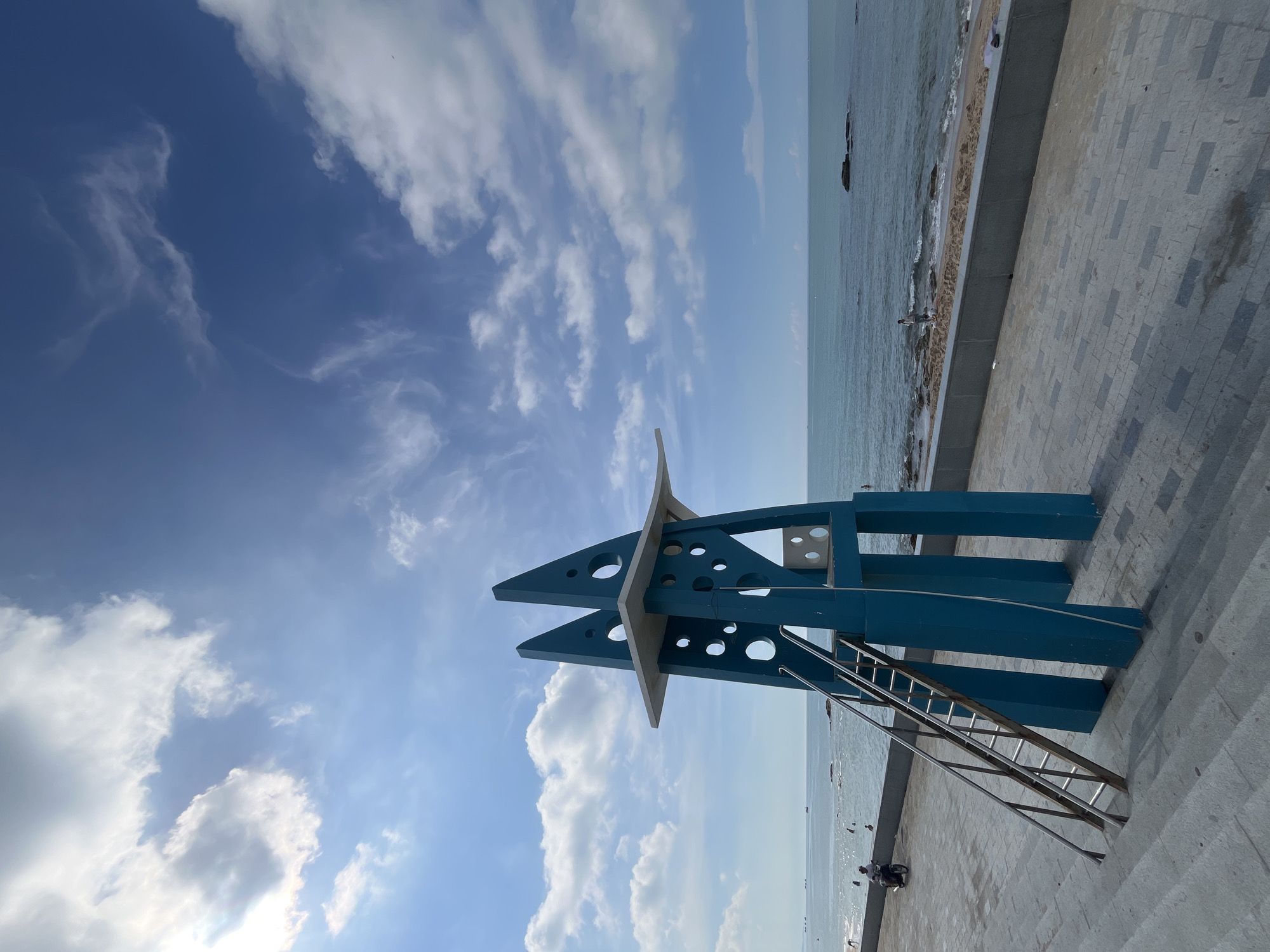 The image depicts a bright, sunny day at a coastal location in Phan Thiết, Vietnam. In the foreground, a striking blue lifeguard tower stands prominently on a paved walkway. The tower has a geometric design with triangular elements and circular cutouts. A metal ladder leans against the tower, leading up to a platform. To the left of the tower, there are concrete stairs.
The walkway extends towards the ocean, where the turquoise water meets a sandy beach. Several people are visible near the water's edge, some wading in the sea. A person is riding a motorcycle on the walkway to the left of the tower, and another individual is standing on the beach in the distance. The sea is calm with a few scattered rocks and distant boats on the horizon. The sky is a vibrant blue, dotted with fluffy white clouds, suggesting fair weather. The overall scene is one of leisure and a pleasant day at the seaside.