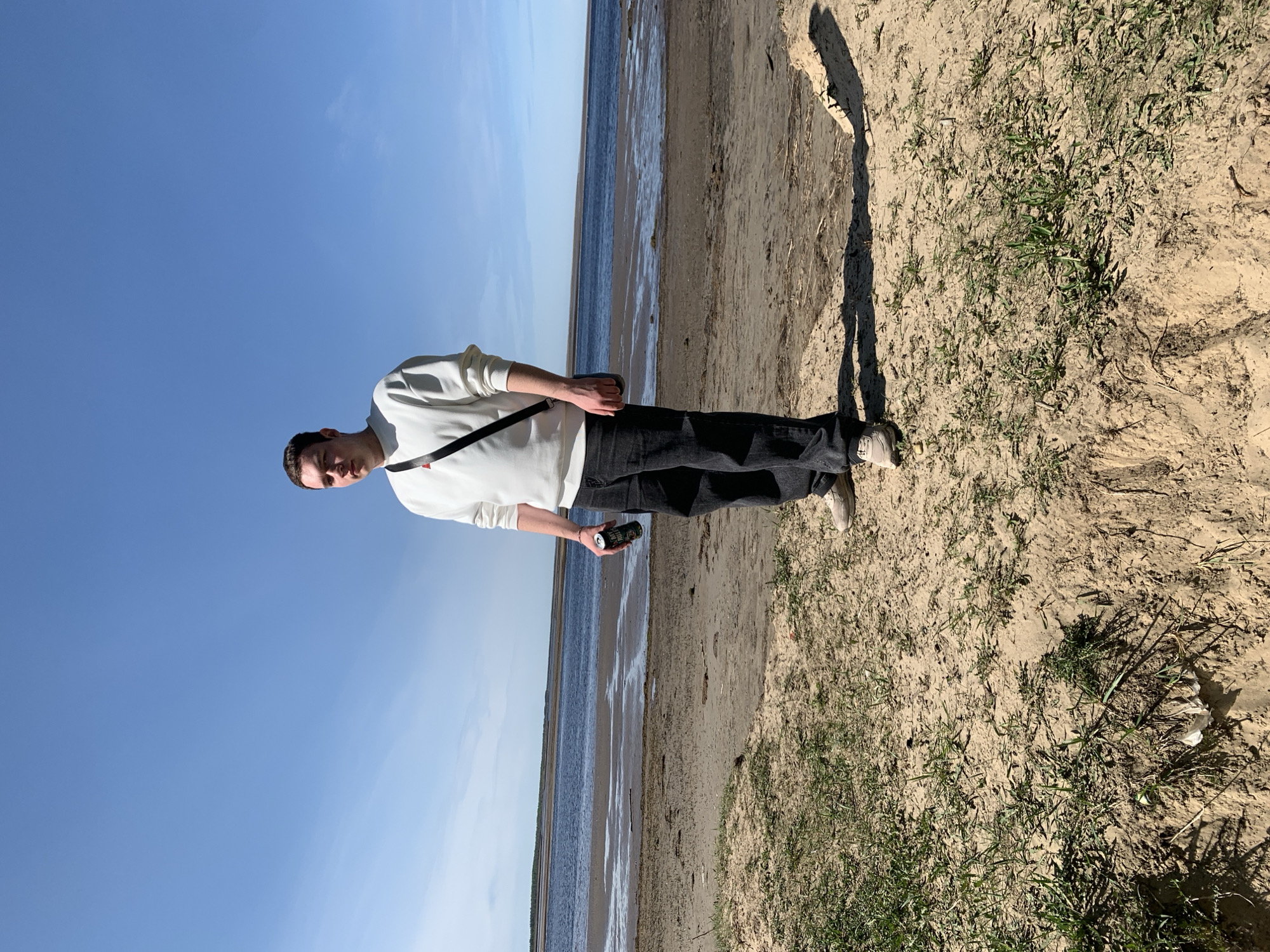 A young man stands on a sandy beach in Severodvinsk, Russia, holding a beverage can. He is wearing a white long-sleeved shirt, dark jeans, and light-colored sneakers, with a black strap across his chest. The background shows a body of water meeting the shoreline under a clear blue sky. Patches of sparse vegetation are scattered across the sand around him.