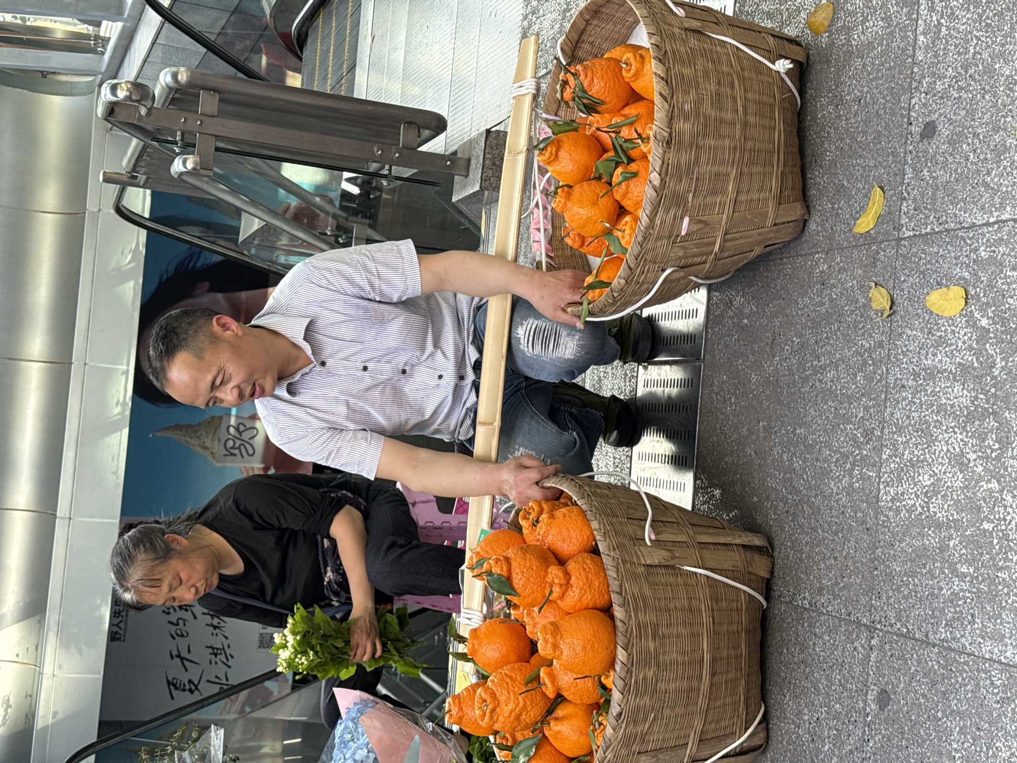 Two individuals sit beside two wicker baskets overflowing with bright orange citrus fruits. The fruits have green leaves still attached. One individual is a man in a striped shirt and jeans, kneeling between the baskets. The other is a woman in a dark shirt. She sits near flowers. They are located near an escalator. The Yuzhong District, China location is inferred from the Chinese writing visible on the glass wall behind the woman. The ground is paved. Several yellow leaves are scattered near one of the baskets.
