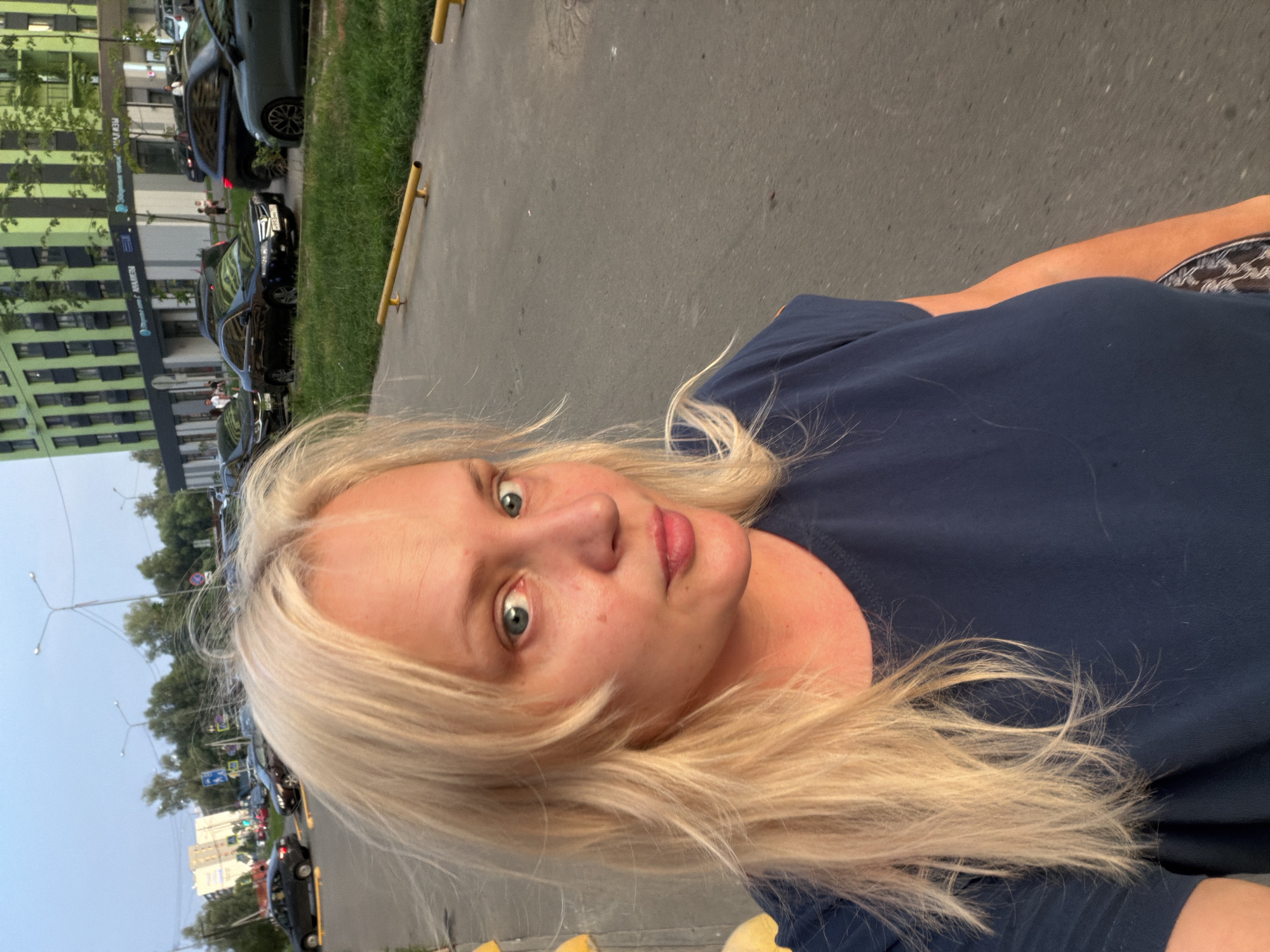 A woman with blonde hair and blue eyes takes a selfie outdoors in Kazan, Russia. She is wearing a dark blue t-shirt and her hair is windswept. In the background, there are parked cars, a green building with windows, and a street light. The woman is standing on a paved surface next to a grassy area with a yellow barrier.