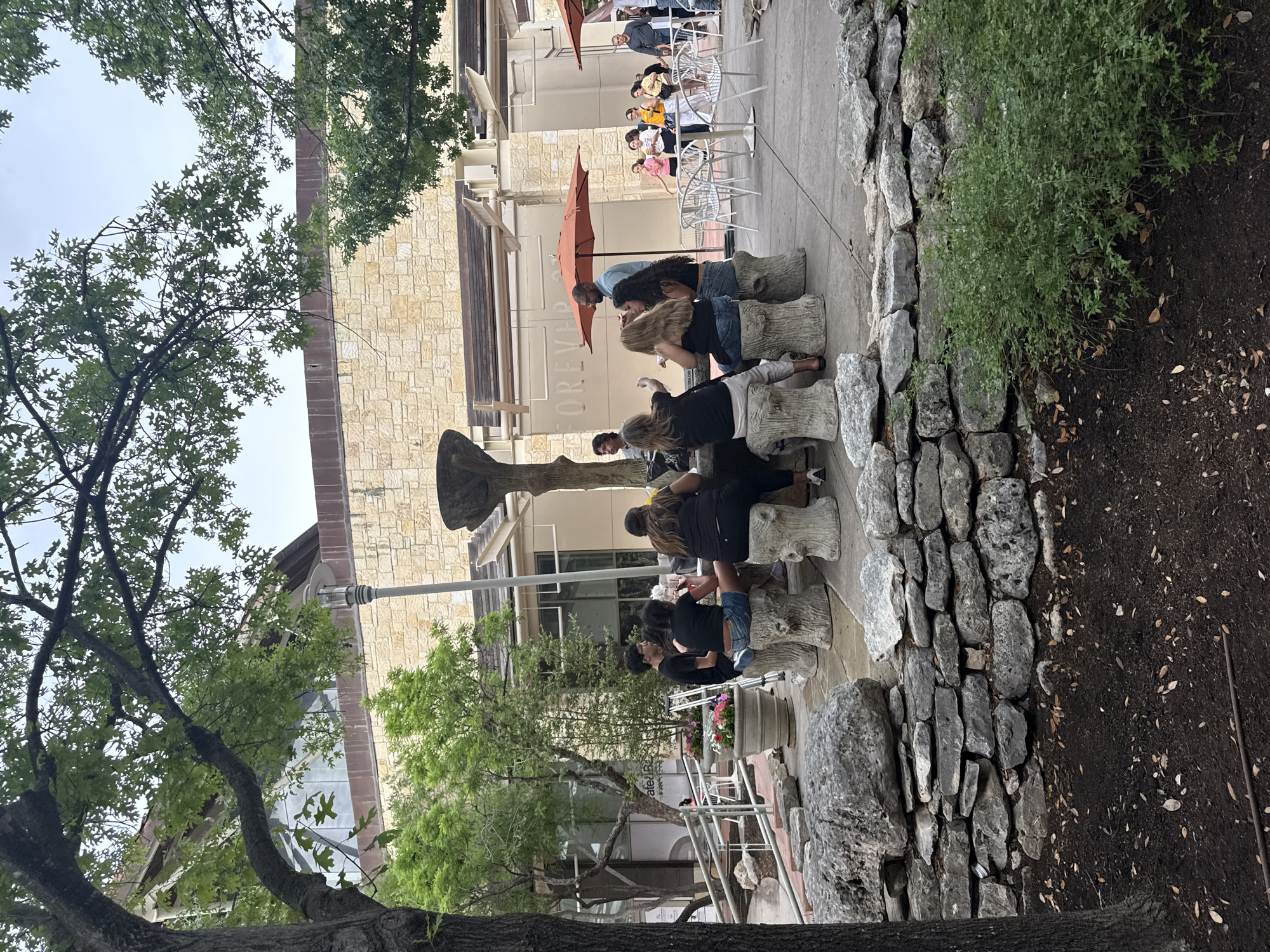 The scene depicts an outdoor area adjacent to a building with the word "FOREVER" visible on its facade, located in San Antonio, United States of America. Several people are sitting on what appear to be faux-stone stools. Additional people are seated at white tables with orange umbrellas in the background. A large, abstract sculpture is visible near the seated group. The area is bordered by a stone wall on one side and is partially shaded by trees.