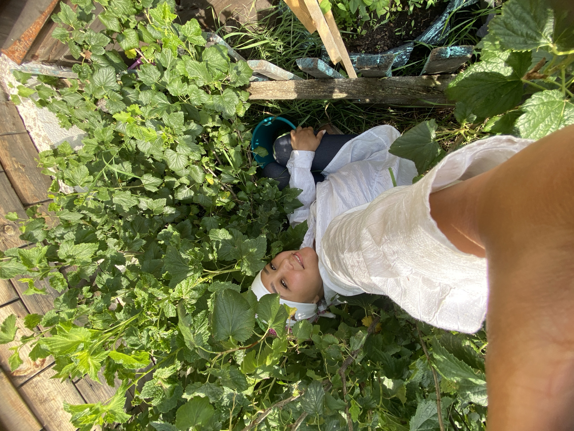 A young woman is lying in a garden, surrounded by lush green bushes. She is wearing a white headscarf and a white shirt. Her eyes are looking at the camera with a smile. A turquoise bucket is visible next to her. The background consists of wooden planks and more greenery, suggesting an outdoor setting in a garden. The scene appears to be a sunny day.