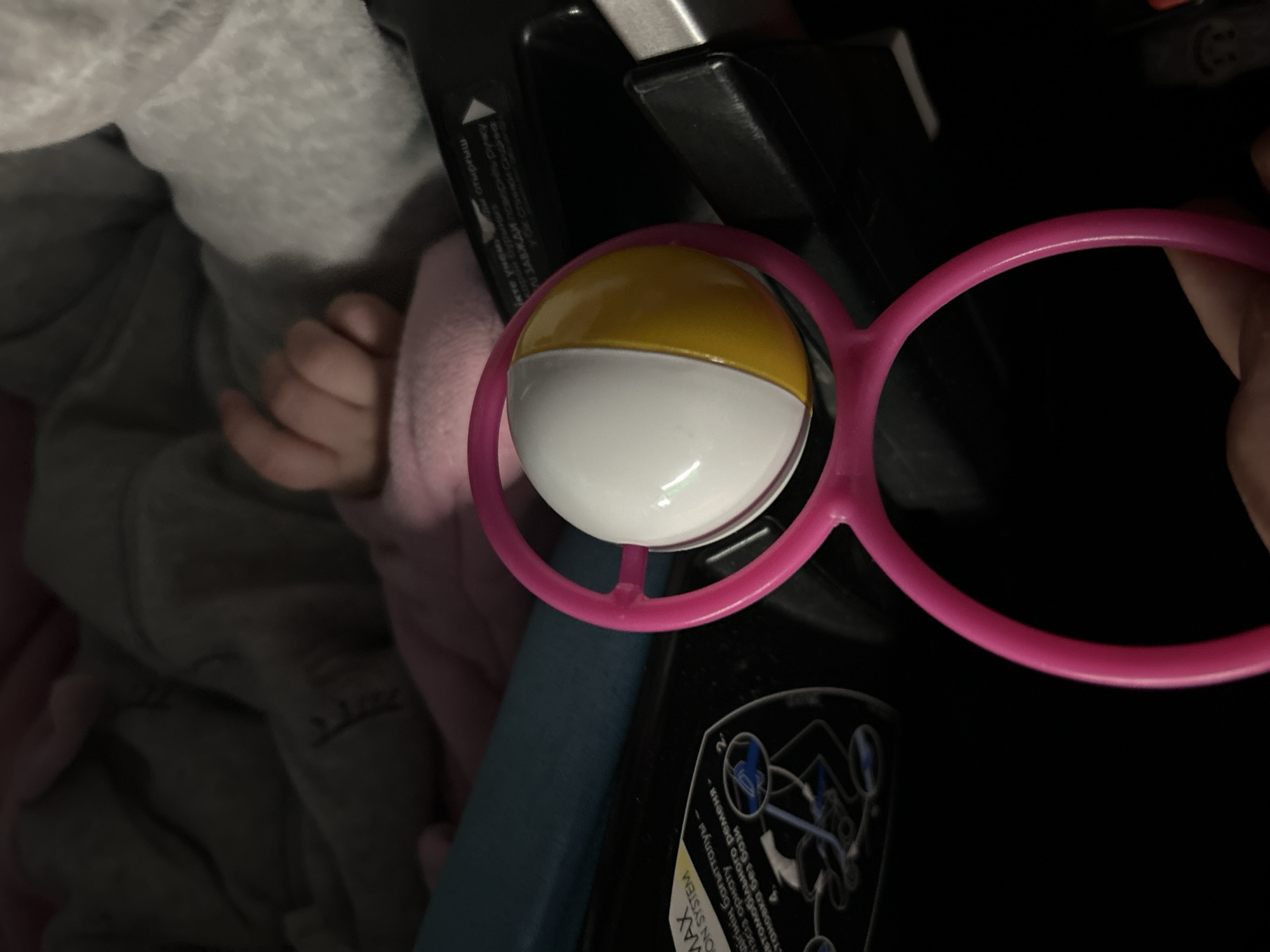 The image shows a baby's hand next to a pink toy rattle with a white and yellow spherical part. The rattle is near what appears to be a car seat, with a portion of the seat's instruction label visible. A person is holding the rattle. There is an unidentifiable portion of clothing to the side of the hand.