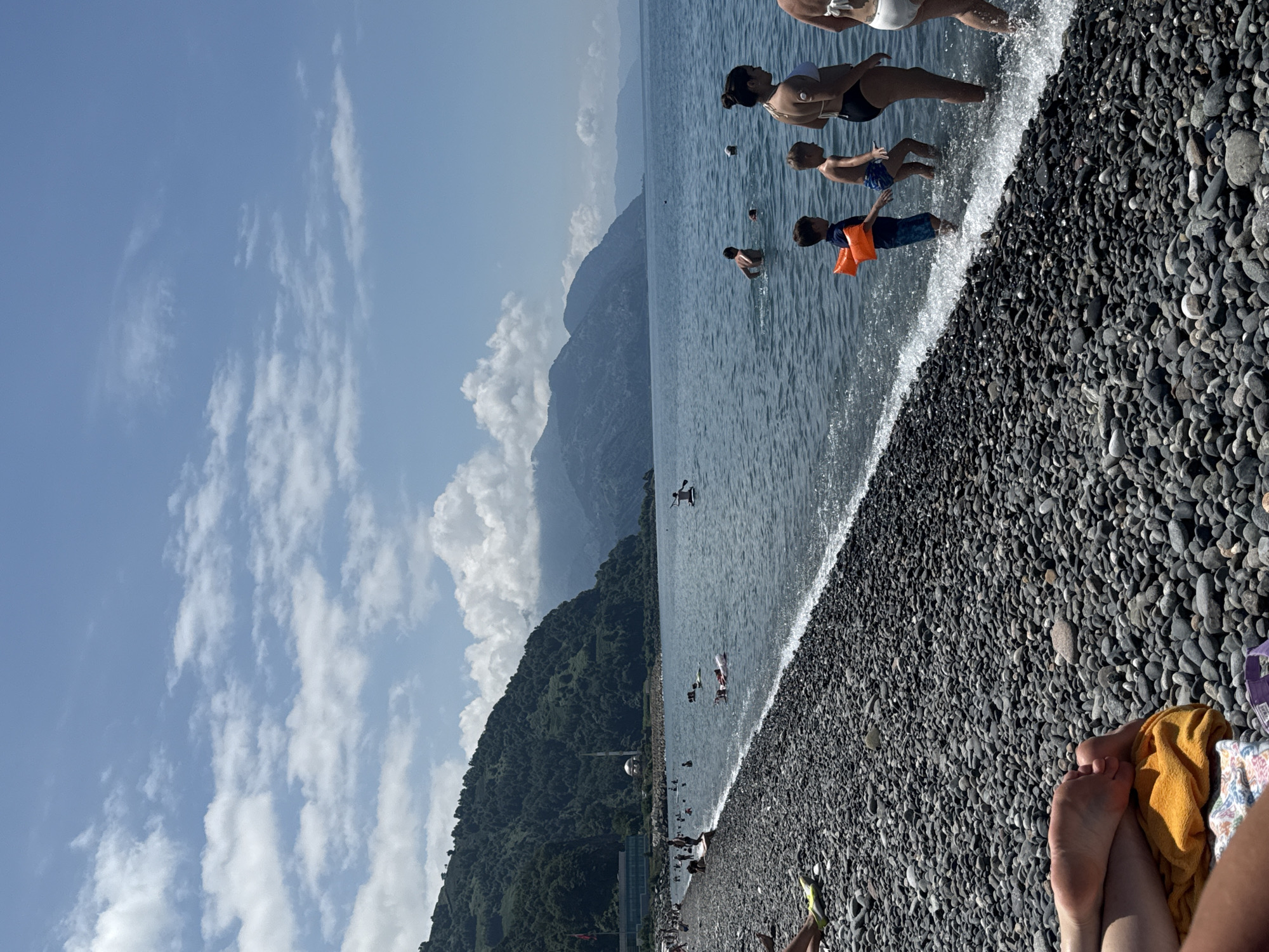 This image depicts a sunny day at a pebble beach in Sarpi, Georgia. The sea is calm and a light blue, with small waves lapping at the shore. In the water, several people are swimming and playing, including children wearing arm floats. A few individuals are standing near the water's edge, while others are further out in the sea.
The beach itself is covered in dark grey and black pebbles. To the left, a lush green, mountainous landscape rises steeply from the coast. A building with a red flag is visible on the hillside, along with a dome structure that appears to be a minaret, suggesting the presence of a mosque. In the distance, more mountains stretch across the horizon under a bright blue sky dotted with white, fluffy clouds.
In the foreground, the legs and feet of a person are visible, resting on a bright orange towel and a patterned blanket. The overall scene is one of leisure and recreation in a scenic coastal environment.