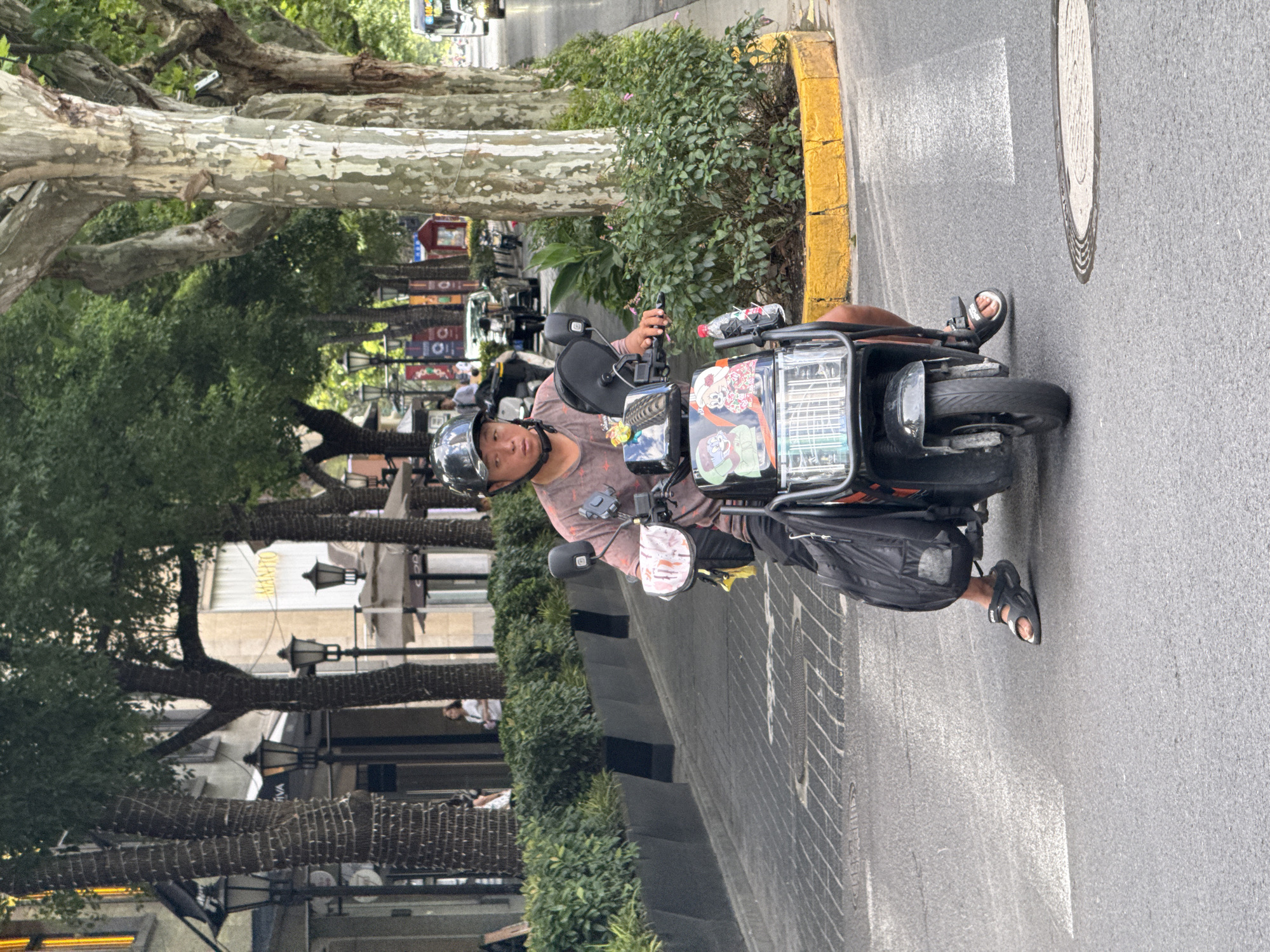 A man wearing a helmet and a patterned t-shirt rides an electric scooter on a paved city street. He is wearing black sandals. The scooter has a black storage bag attached to the back and decorative stickers on the front panel. He is holding the handlebars with his right hand. The scooter has a license plate that is not visible. In the background, there are trees with mottled bark lining the street, buildings with awnings, and streetlights. A yellow curb separates the street from a planted area with green bushes. The sky is overcast, and the lighting suggests it is daytime. The scene appears to be a street in Shanghai, China, given the architecture and streetscape typical of the Bund area.