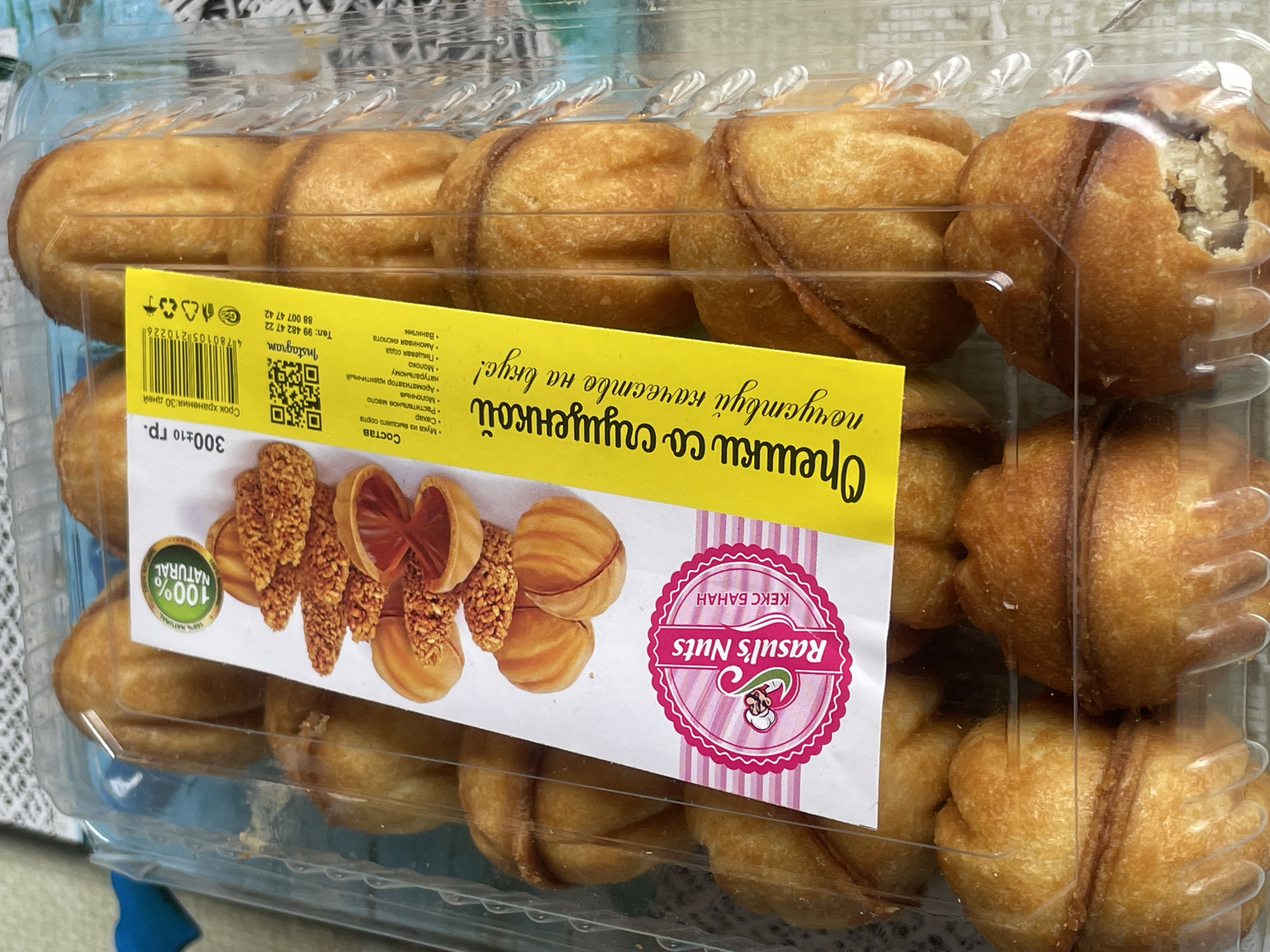 The image shows a clear plastic container filled with golden-brown baked goods, resembling small, hollowed-out cookies or pastries. The container is partially filled, with multiple layers of these pastries visible. A yellow label is affixed to the front of the container, featuring text in a Cyrillic script and some graphics. The label includes a circular emblem with "100% NATURAL" written on it, as well as images of ingredients or finished products, including what appears to be a split pastry filled with a reddish-brown substance and crushed nuts. A brand name "Rasul's Nuts" is also prominent on the label. The background of the image is out of focus but suggests an indoor setting with a light-colored, possibly textured surface. The overall impression is of a commercial product for sale, likely in a bakery or supermarket. There is no discernible information in the image to indicate the location as Tashkent, Uzbekistan.