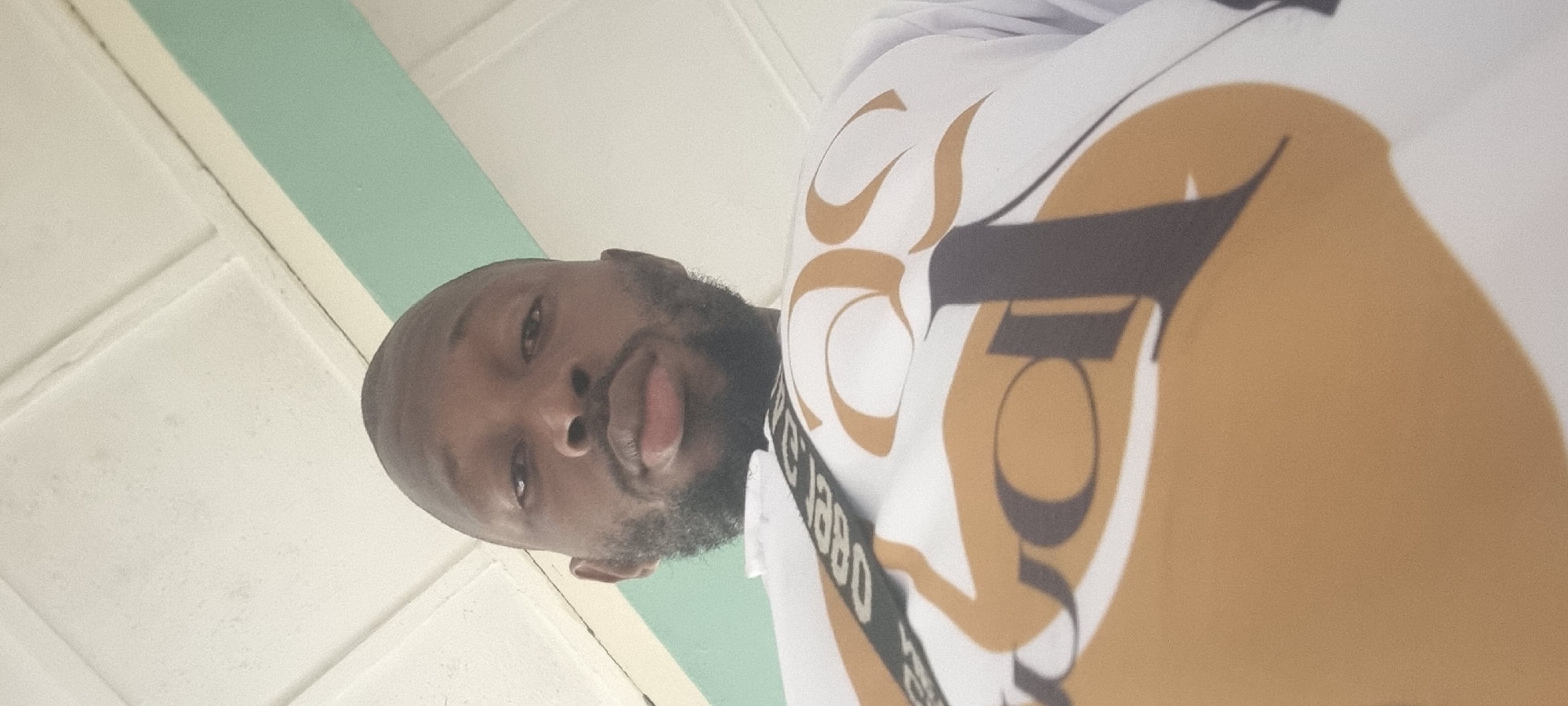 A medium shot from a lower angle captures a bearded man with a shaved head, wearing a white shirt with a large, stylized, tan and brown design. The man appears to be looking down at the camera. Behind him and slightly to the left, a white ceiling with a pale green stripe is visible. The location is in the city of Unknown, Unknown.