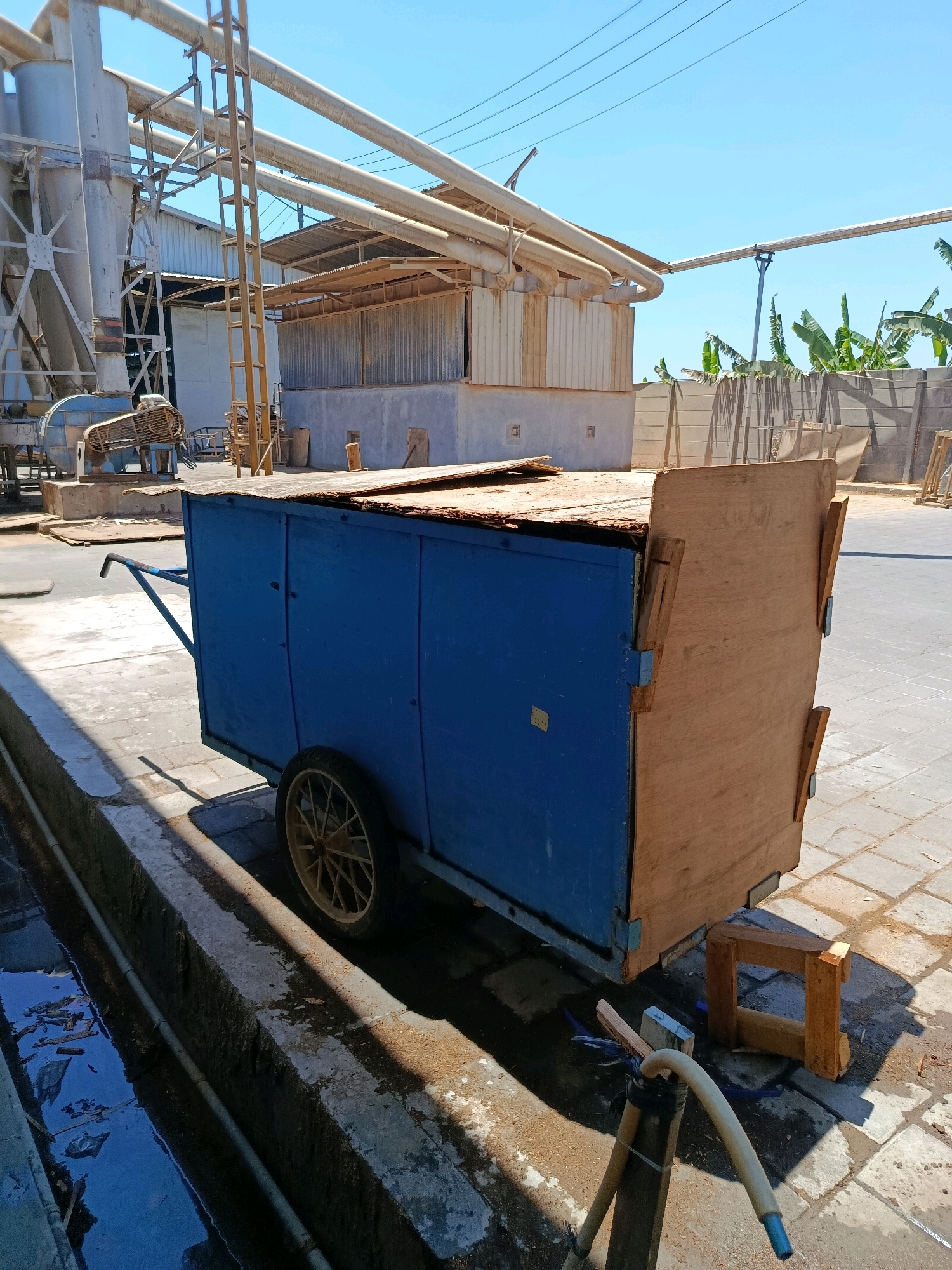 In Kembangarum, Indonesia, an outdoor industrial setting is depicted under a clear blue sky. The foreground features a blue, wooden cart with a single spoked wheel. The cart's wooden top appears weathered and partially dislodged. To the left, a concrete channel filled with dark, murky water runs alongside the cart. In the background, industrial structures include large, grey cylindrical silos and a network of pipes overhead. A fan or blower unit is visible attached to one of the silos. The ground is paved with concrete tiles. Banana trees are visible in the distance, suggesting a tropical climate. The scene appears to be a functional area within an industrial complex.