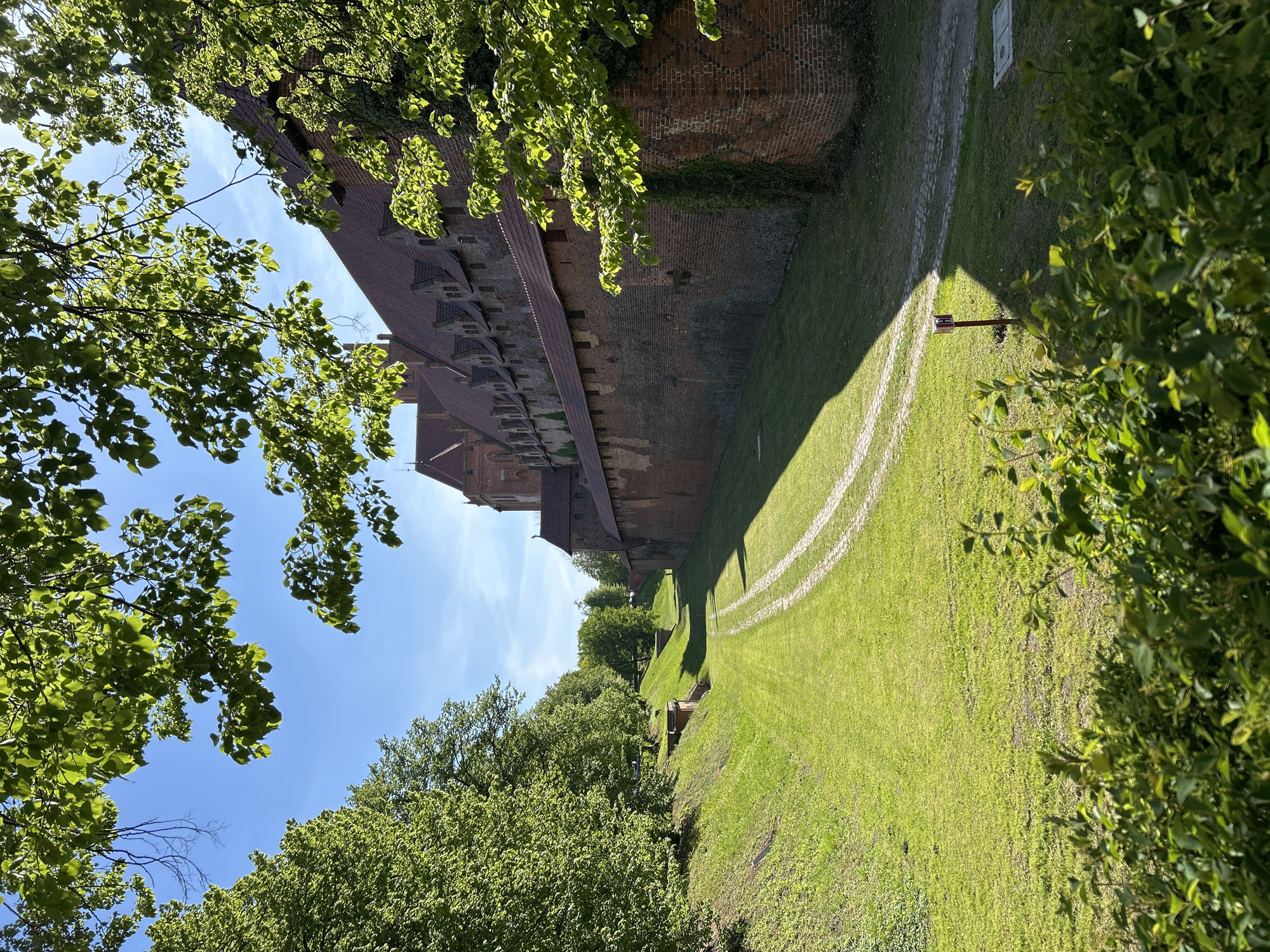 The image depicts a section of the Malbork Castle moat in Malbork, Poland. A tall, brick castle wall lines one side of the frame, partially obscured by leafy trees in the foreground. A wide, grassy moat stretches beside the wall, marked with white painted lines. More trees line the far side of the moat. The sky is visible, with patches of blue and scattered clouds.