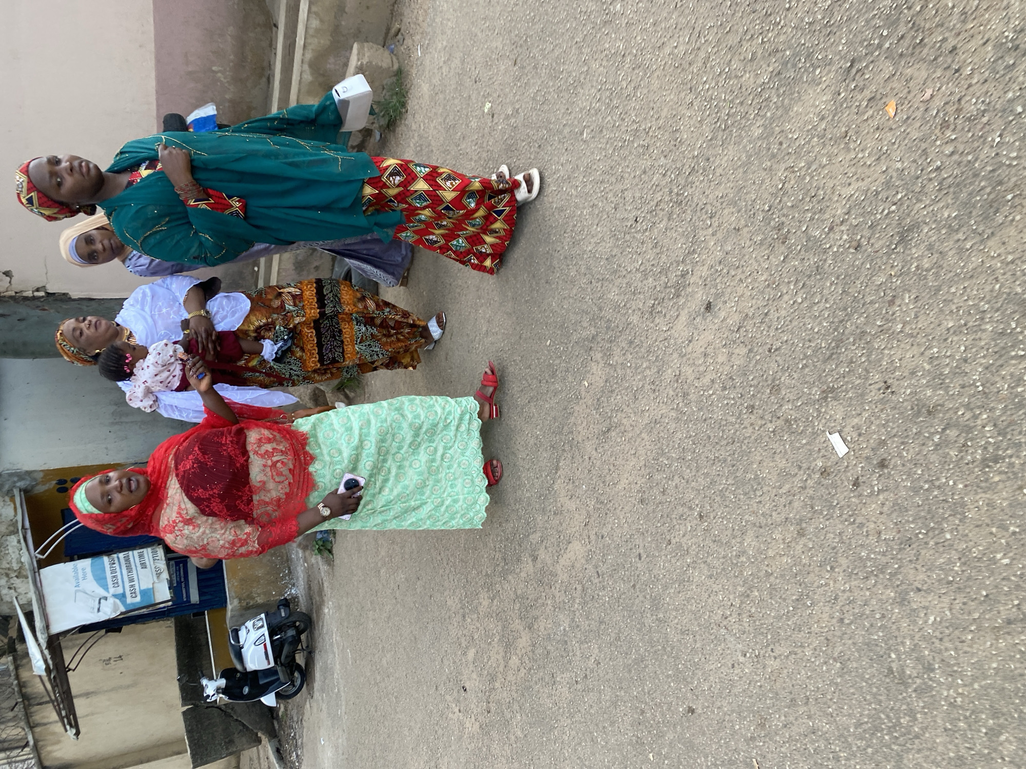 A group of women and a child are walking on a paved street in Jos, Nigeria. The woman in the foreground on the left is wearing a red headscarf, a red and beige patterned top, and a light green patterned skirt. She is holding a smartphone in her right hand and wearing red sandals. To her right, a woman in a white top and patterned skirt is holding a baby wearing a light pink and white patterned outfit. Behind them, a woman in a grey veil and patterned skirt is walking, followed by a woman in a teal headscarf and teal garment with gold trim, and a patterned red and yellow skirt. She is holding a white bag in her right hand and wearing white sandals. In the background, a white scooter is parked next to a kiosk with a sign displaying services like "Available Here", "Cash Deposit", "Cash Withdrawal", and "Airtime". The environment appears to be an urban street with a building and a concrete barrier.