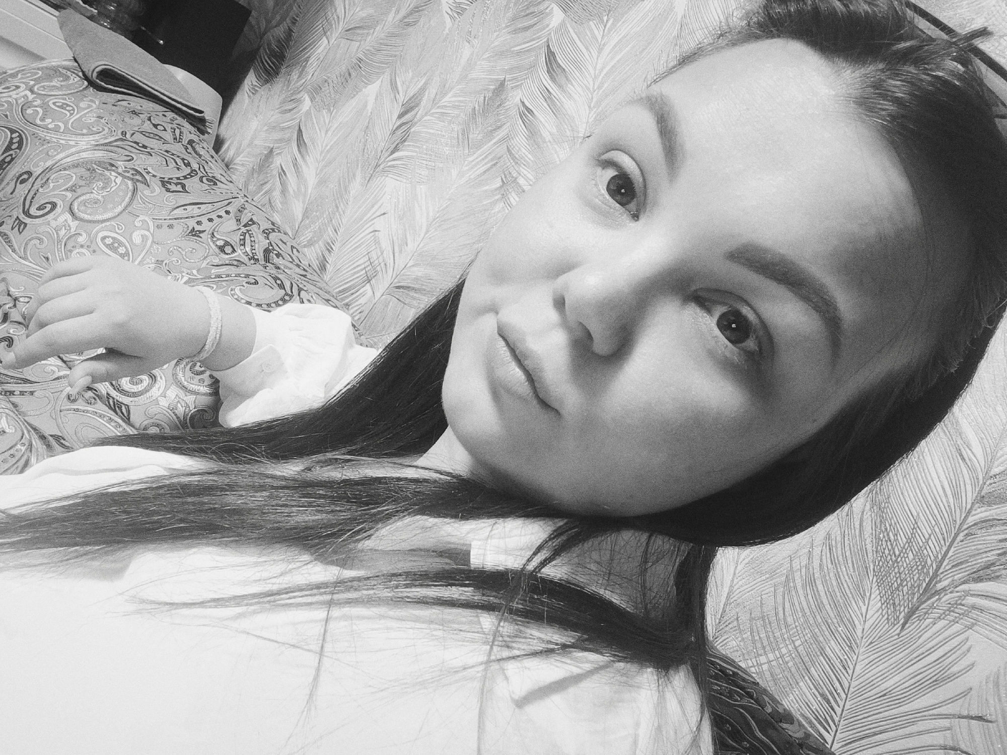 A black and white selfie shows a young woman lying down. She has long, dark hair and is wearing a light-colored shirt. She is looking at the camera with a slight smile. Her right hand rests on a pillow with a paisley pattern. The background is a wall with a feather-like pattern.
There is no evidence of location in the city of Прикамский, Russia.