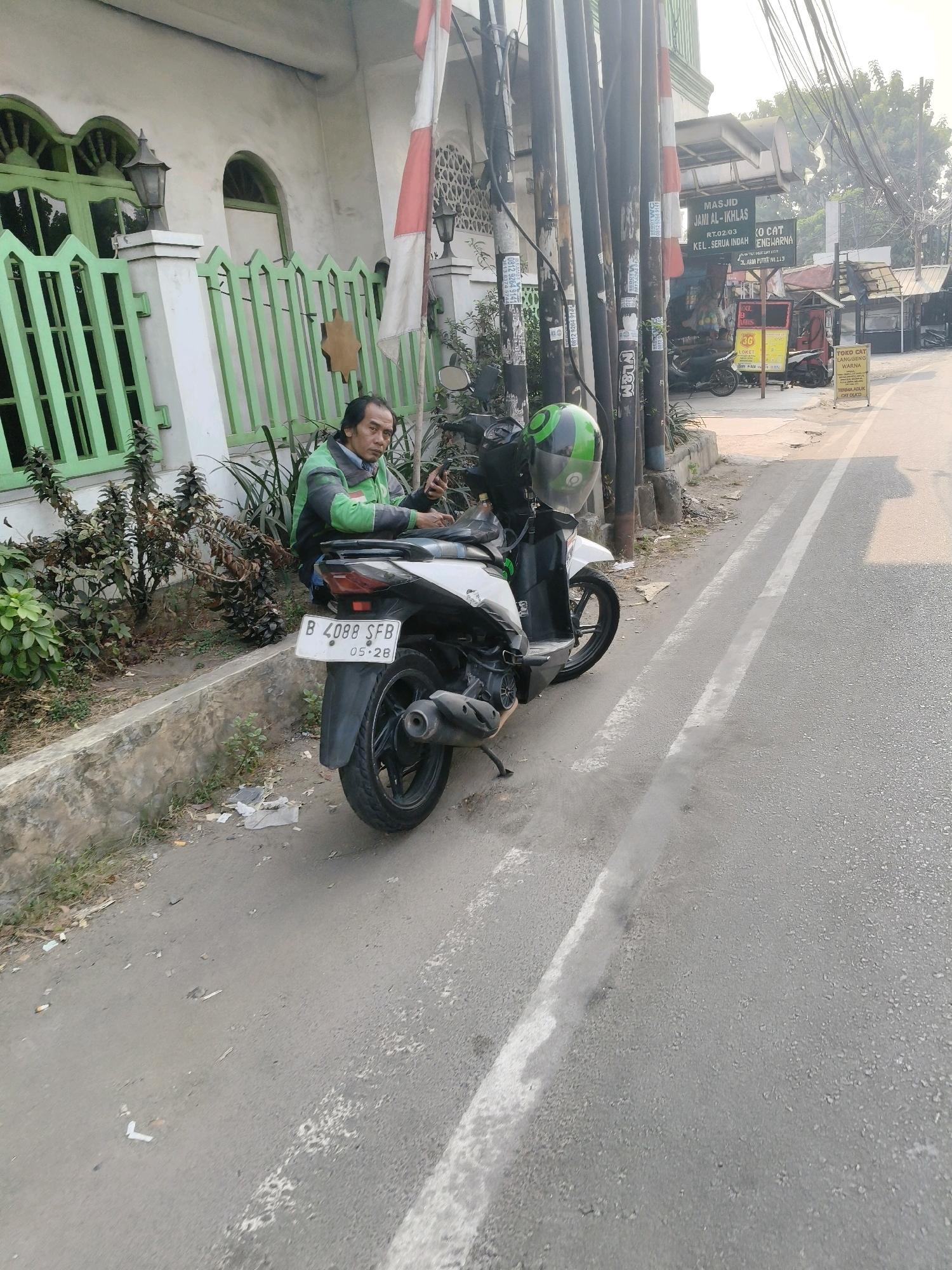 A man wearing a green and black jacket and blue jeans is sitting on a motorcycle on the side of a road in South Tangerang, Indonesia. He is looking at his phone. The motorcycle is black and white. Behind the man is a green fence and a building. In the background, there are shops and a mosque. The road is asphalt and there are white lines marking the lanes. The sky is overcast.