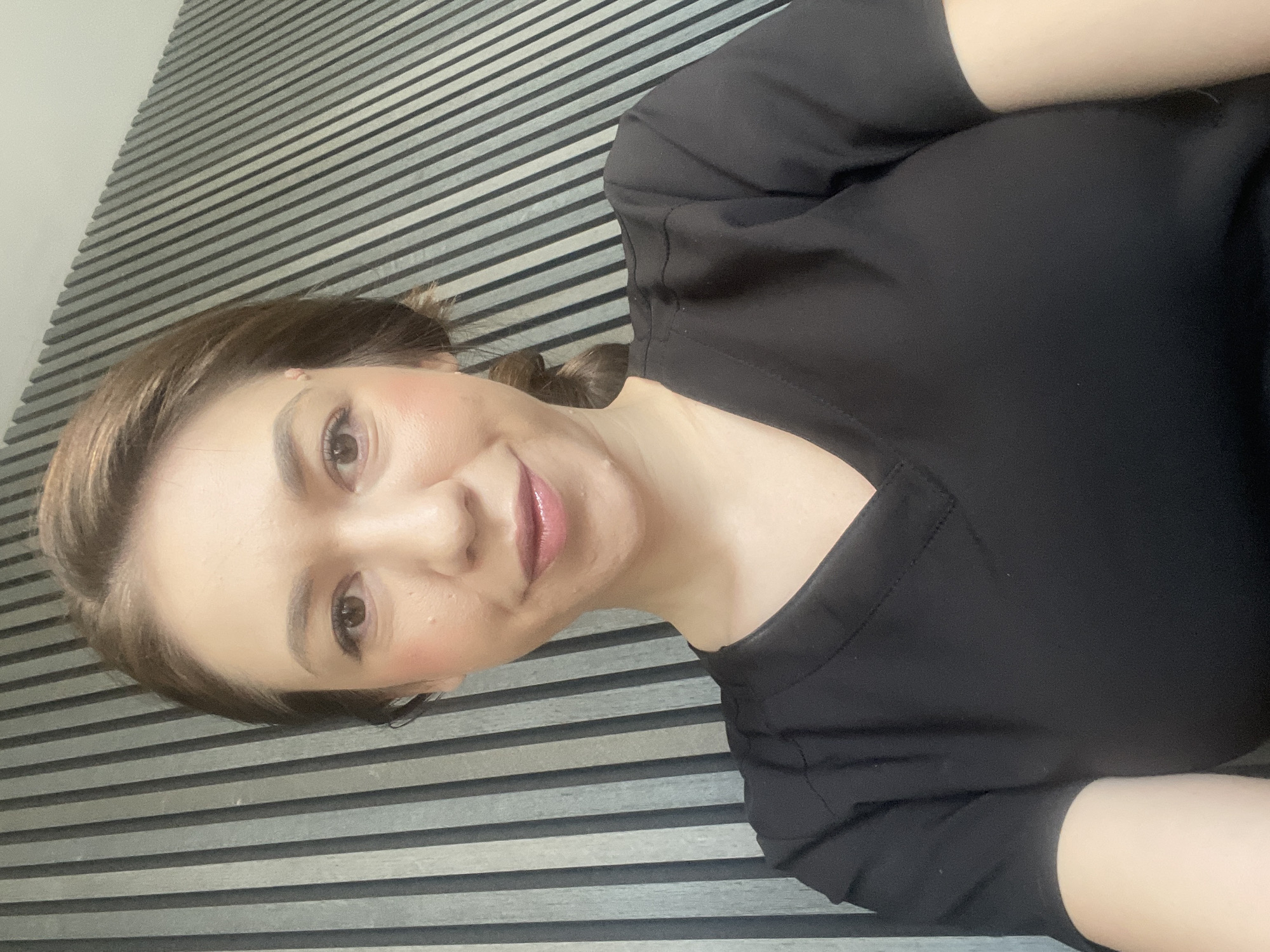A woman with light brown hair and fair skin is lying down on a surface with horizontal, alternating gray and white stripes. She is wearing a black, V-neck top. Her head is tilted, and she is looking at the camera with a slight smile. Her makeup is noticeable, with blush on her cheeks and lipstick. The background consists solely of the striped surface. There is no indication of location in Sofia, Bulgaria, or any other specific city. The image focuses solely on the woman and the striped surface.