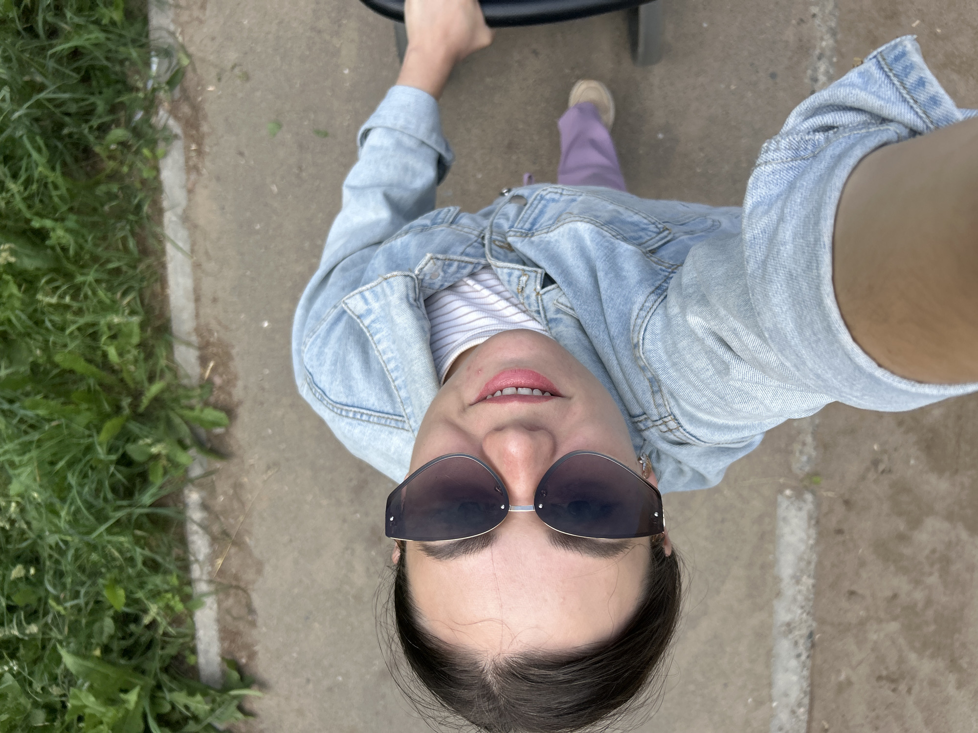 A high-angle selfie shows a young woman from the shoulders up, with her face turned towards the camera. She is wearing a light blue denim jacket over a striped white t-shirt, lavender pants, and tan shoes. Large, dark-lensed sunglasses obscure her eyes, and her lips are parted to reveal white teeth and pink lipstick. She has dark hair pulled back. Her right hand is visible holding onto a black object. The background consists of a grey concrete path bordered by a strip of green grass and vegetation. The scene appears to be outdoors, possibly in a park or along a walkway in the city of Chaikovsky, Russia.