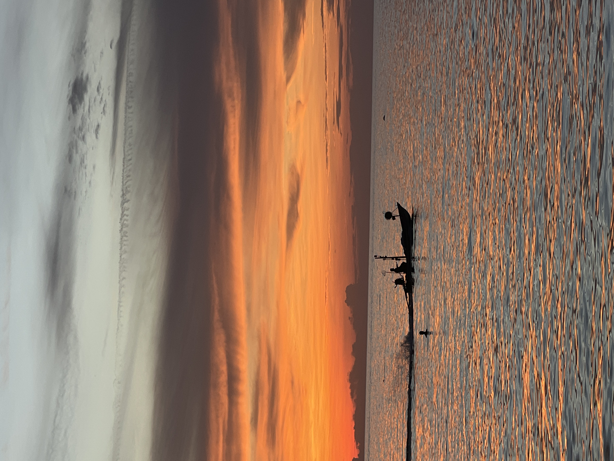 The image captures a serene sunset scene over the ocean in Ko Pha-ngan District, Thailand. The sky is painted with vibrant hues of orange and yellow, transitioning into darker grays and pale blues towards the top. Wispy clouds are scattered across the sky, with some appearing as faint streaks.
In the water, a small fishing boat is silhouetted against the colorful sky. The boat appears to be moving, creating a wake behind it. Two figures are visible on the boat, one in the front and another towards the back. One figure seems to be holding a net. There is also a single person silhouetted in the water, appearing to be swimming or floating.
The ocean surface reflects the colors of the sunset, creating a shimmering, textured effect. The horizon line is visible, separating the sea from the sky. The overall impression is one of tranquility and natural beauty.