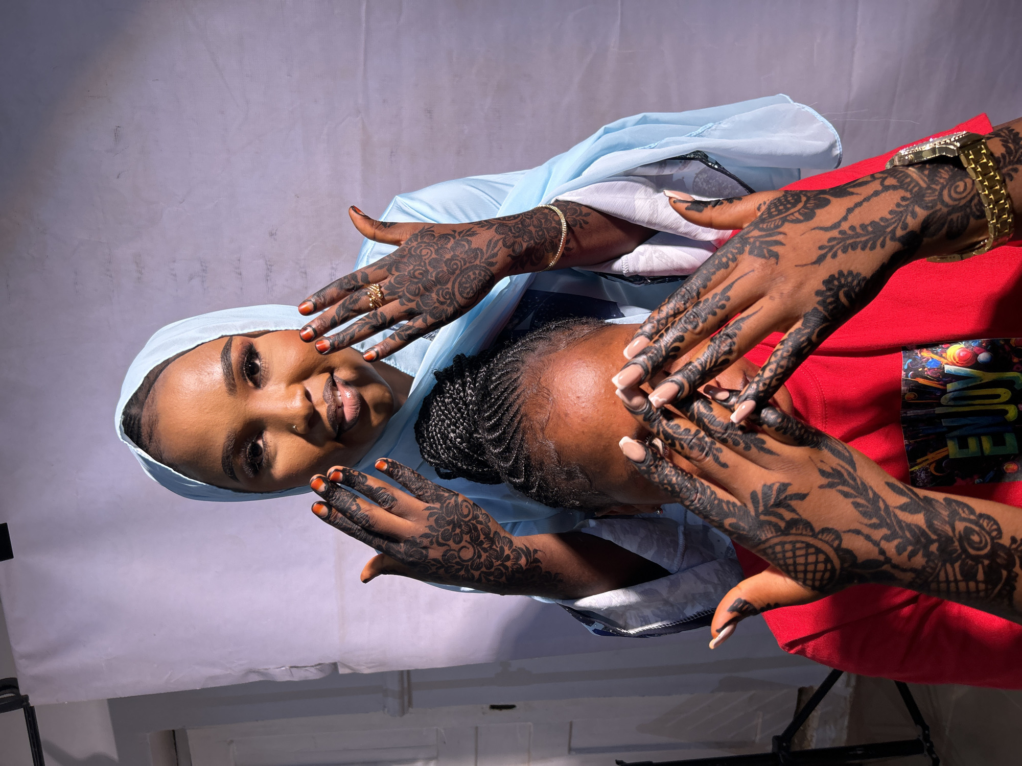 Two women are pictured in an indoor setting. One woman wears a light blue headscarf and a matching shawl. Her hands, adorned with henna designs, are positioned near the face of the second woman. The second woman wears a red garment. Her hands also feature henna designs and are placed near the first woman's face. The photograph appears to be taken indoors. There is no indication that it was taken in the city of Jos, Nigeria.