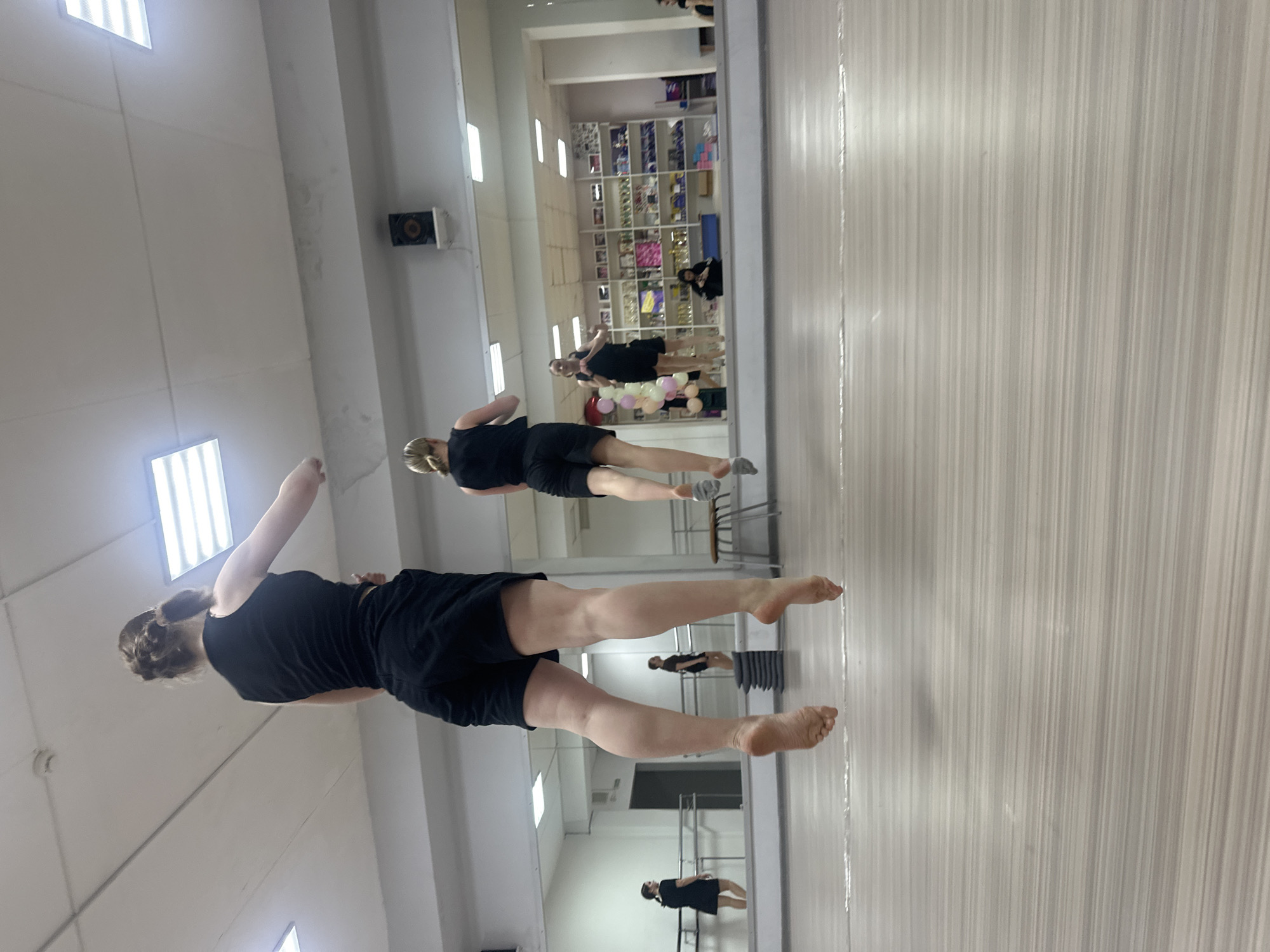 The image depicts a woman in a black t-shirt and shorts, with her hair in a bun, seemingly mid-movement in a dance studio. She is barefoot and her body is positioned in a way that suggests motion or dance.
The studio has a large mirror covering one wall, reflecting the woman and the studio space. Inside the mirror's reflection, one can see additional people, along with what appears to be a wall of shelves stocked with various items.
The location may not be possible to determine based on the current information available in the image. Without specifics tied to location the city of Votkinsk, Russia may not be accurate.
