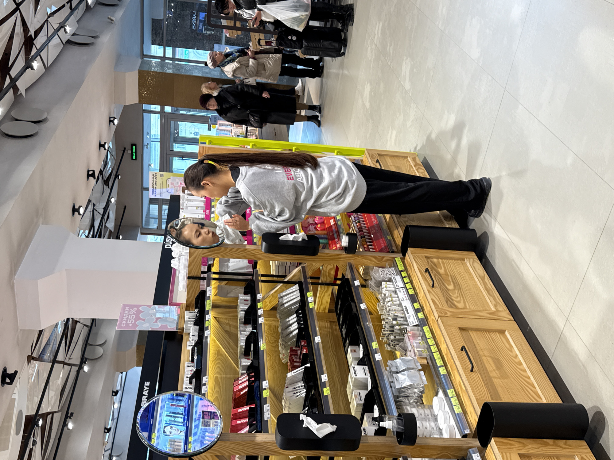 The image captures a scene inside a brightly lit beauty store or cosmetics department in Kazan, Russia, during the daytime. In the foreground, a young woman with long dark hair, tied back with a yellow scrunchie, wears a grey hooded sweatshirt with partial text "EVER ABO" and black pants. She leans over a wooden display counter, intently looking at her reflection in a circular mirror, appearing to examine her face or apply makeup.
The wooden display is well-stocked with various beauty products including lipsticks, creams, and packaged items, with visible brand names like "AVE", "BAYE", "RAVE", and "BRAVE". Black tissue box holders containing white tissues and a hand sanitizer dispenser are also placed on the counter. A pink sign above the display advertises "скидки -55%" (discounts -55%). In the background, several other customers are out of focus, appearing to queue or browse. They are dressed in coats, suggesting cooler outdoor temperatures. A digital display in the background shows "ПОДАРОЧНАЯ КАРТА ОНЛАЙН" (Gift Card Online), and through large glass doors, a green "BЫХОД" (EXIT) sign is visible. The store features a light-colored tiled floor and modern overhead lighting.