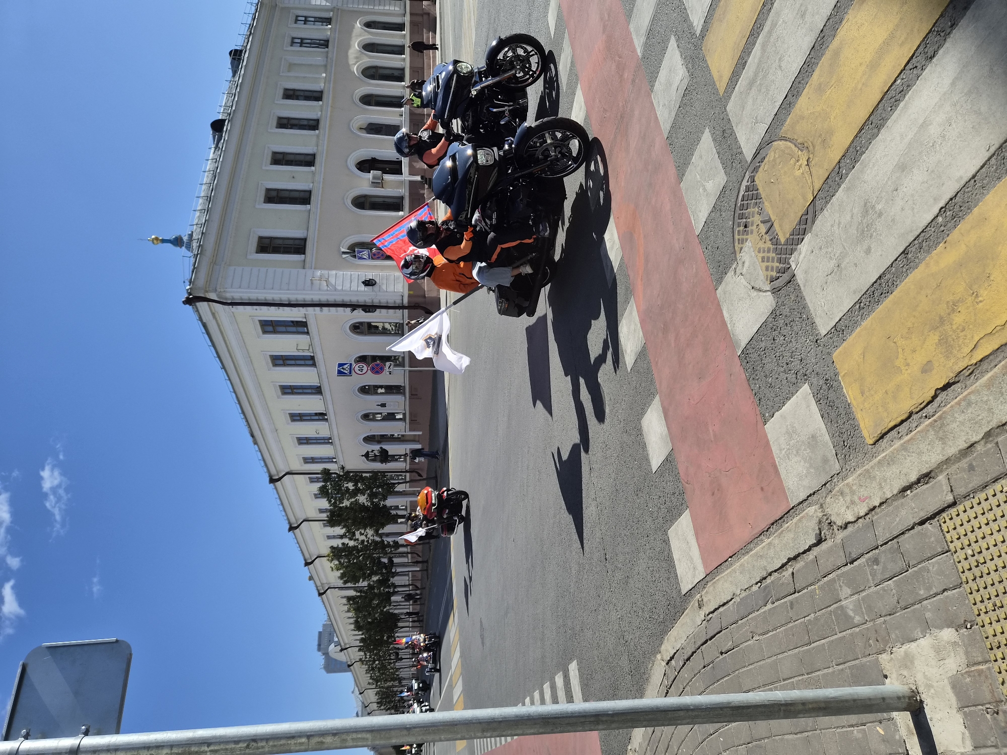 This is a wide shot of a parade with motorcycles in Kazan, Russia, on a sunny day. In the foreground, a street with a pedestrian crossing is visible, with red and yellow stripes. A metal pole stands on the left side of the image. Several motorcycles are seen riding in a line on the street. The first motorcycle in the foreground has two people on it, the rider and a passenger. Both are wearing helmets and orange vests. The passenger is holding a white flag. Behind this motorcycle, another motorcycle is visible, also with two people. They are wearing helmets and orange vests, and the passenger is holding a red and blue flag. Further down the street, more motorcycles with riders and passengers are seen, some with flags. The background shows a long, light-colored building with arched windows, possibly a historical building, and a clear blue sky with a few white clouds. Some trees line the street in front of the building. There are traffic signs visible on the side of the road. The scene appears to be taking place on a city street during a sunny day.