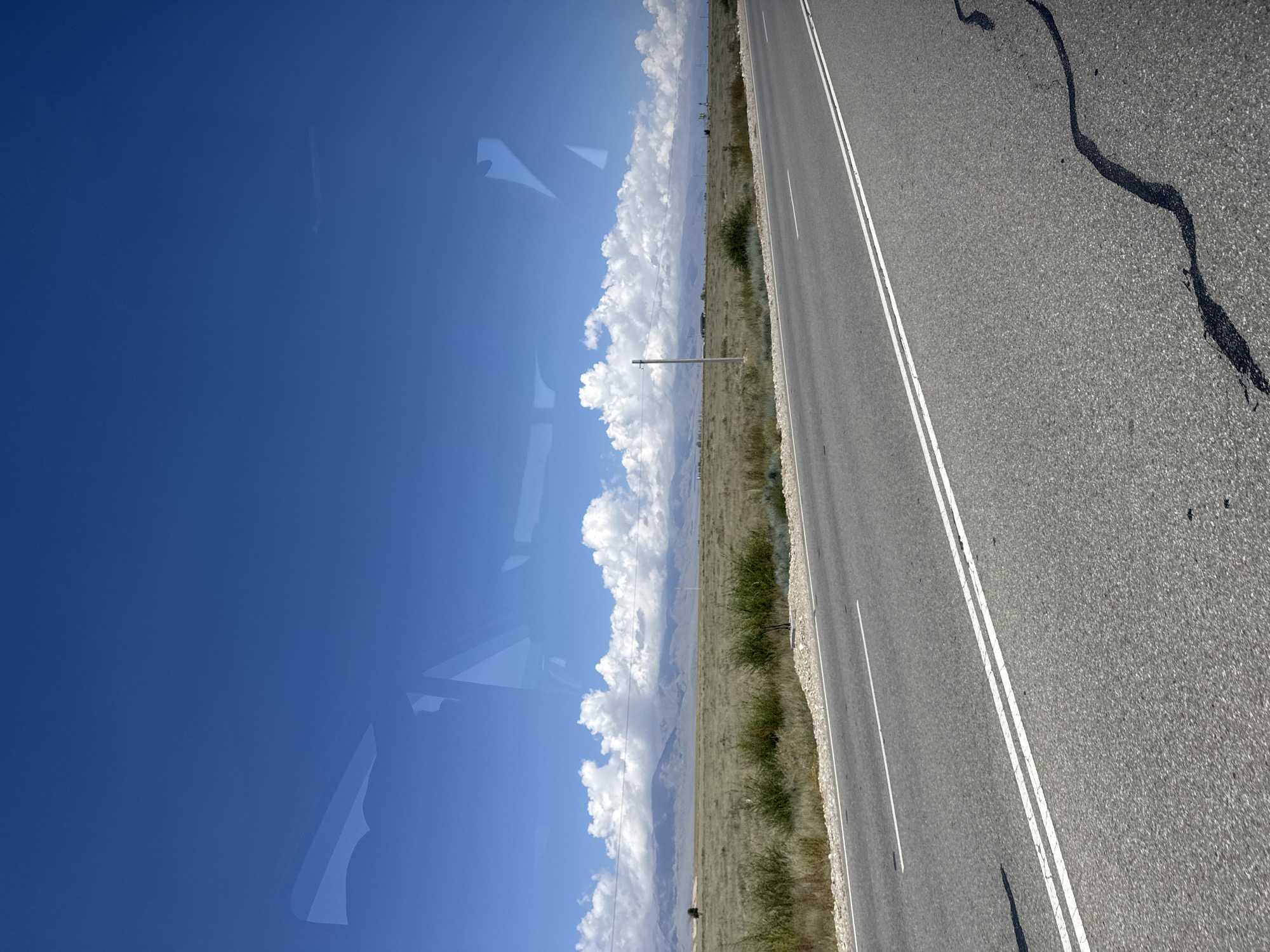 The image depicts a roadside view of a paved road with double white lines, leading towards a vast, dry, grassy landscape under a bright blue sky dotted with fluffy white clouds. In the distance, a mountain range is visible, partially obscured by clouds. A single utility pole stands in the middle ground. Reflections of what appear to be vehicle windows are visible in the upper part of the image. The scene is serene and expansive, typical of rural or semi-arid environments. The location is identified as Toru-Aigyr, Kyrgyzstan.