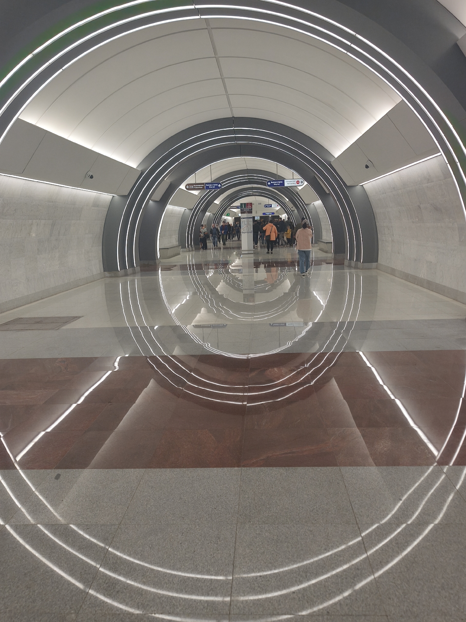 A wide, angled shot of a subway station concourse with numerous passengers in the distance. The concourse is defined by a curved, tunnel-like structure with multiple concentric rings of light extending from the ceiling. These illuminated rings are reflected on the polished floor. The floor is divided into two distinct sections, a light gray section and a maroon section. The station signage is visible in the distance, and security cameras are mounted near the ceiling on the left side. The lighting is bright and modern.