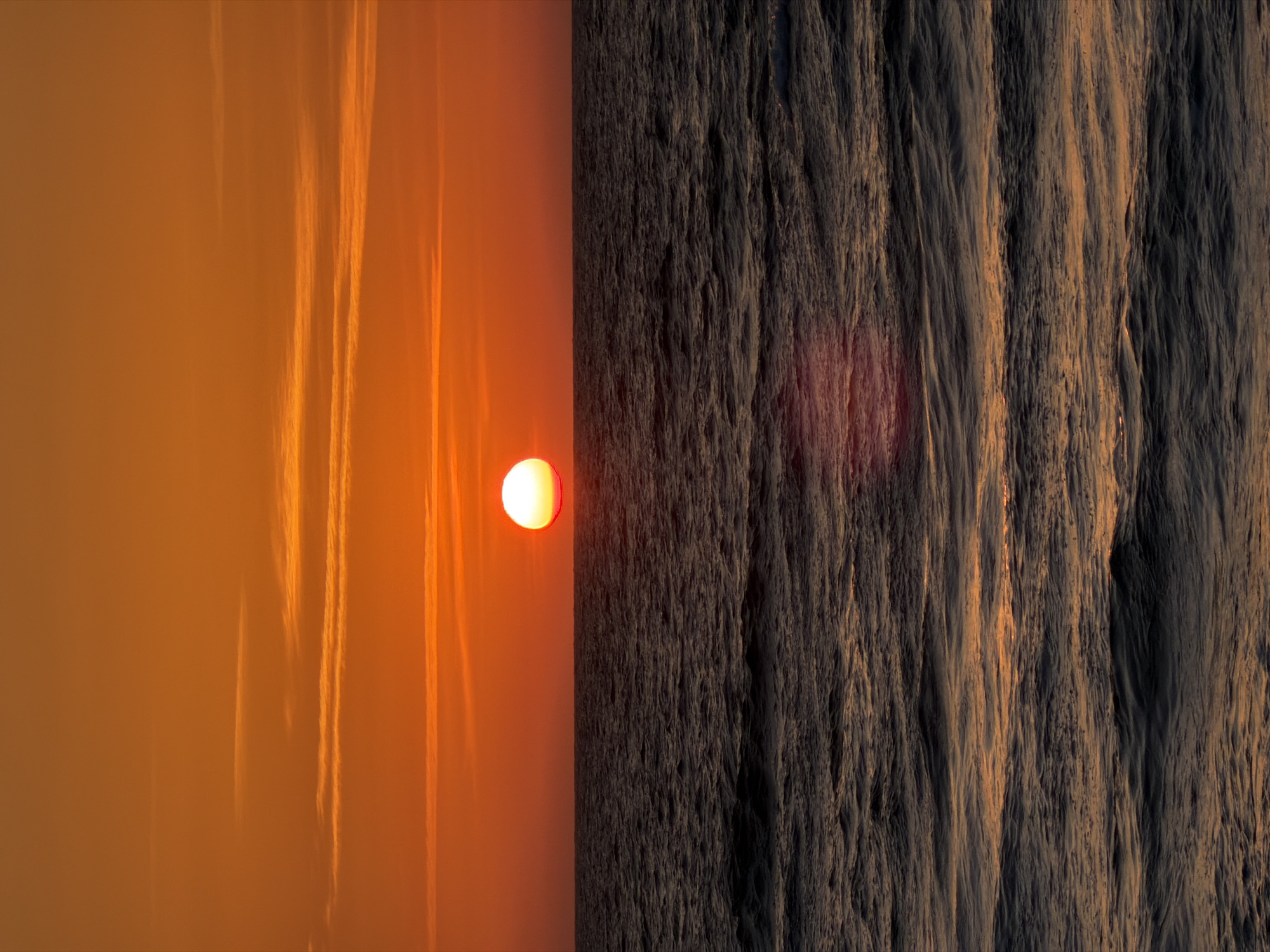 An eye-level, outdoor shot depicts a sunrise or sunset in Tel Aviv, Israel, with the image oriented sideways. The sky occupies the left half of the frame and is a gradient of orange hues with blurred streaks suggesting cloud formations or condensation trails. A bright, circular sun is positioned near the center, casting a warm glow. The right half of the image features the Mediterranean Sea, its surface textured with ripples and waves that catch the light, rendering the water in shades of gray and dark brown. The lack of sharp detail suggests either low lighting or a focus on the broad atmospheric effects. No people, specific objects, or actions are discernible in the photograph.