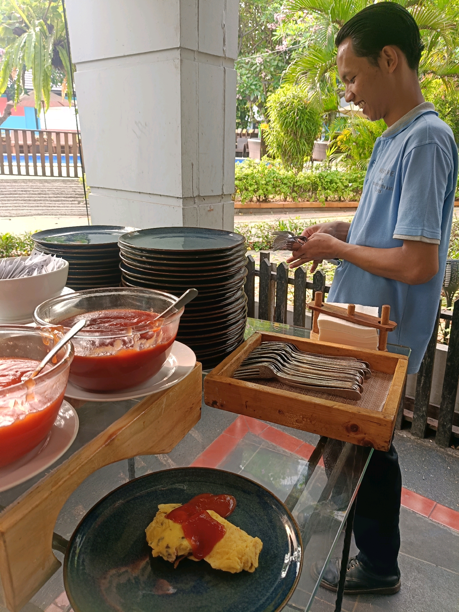 A man wearing a light blue polo shirt and black pants is standing at a buffet table in North Jakarta, Indonesia. He is smiling and holding a stack of forks in his hands. In front of him is a wooden tray filled with more forks, a wooden napkin holder with white napkins, and two glass bowls filled with a red sauce. To the left of the bowls are stacks of dark blue plates. On a dark blue plate in the foreground, there is a piece of food covered in the red sauce. The buffet table is outdoors, with lush green foliage and trees in the background. The man appears to be preparing the buffet for guests.