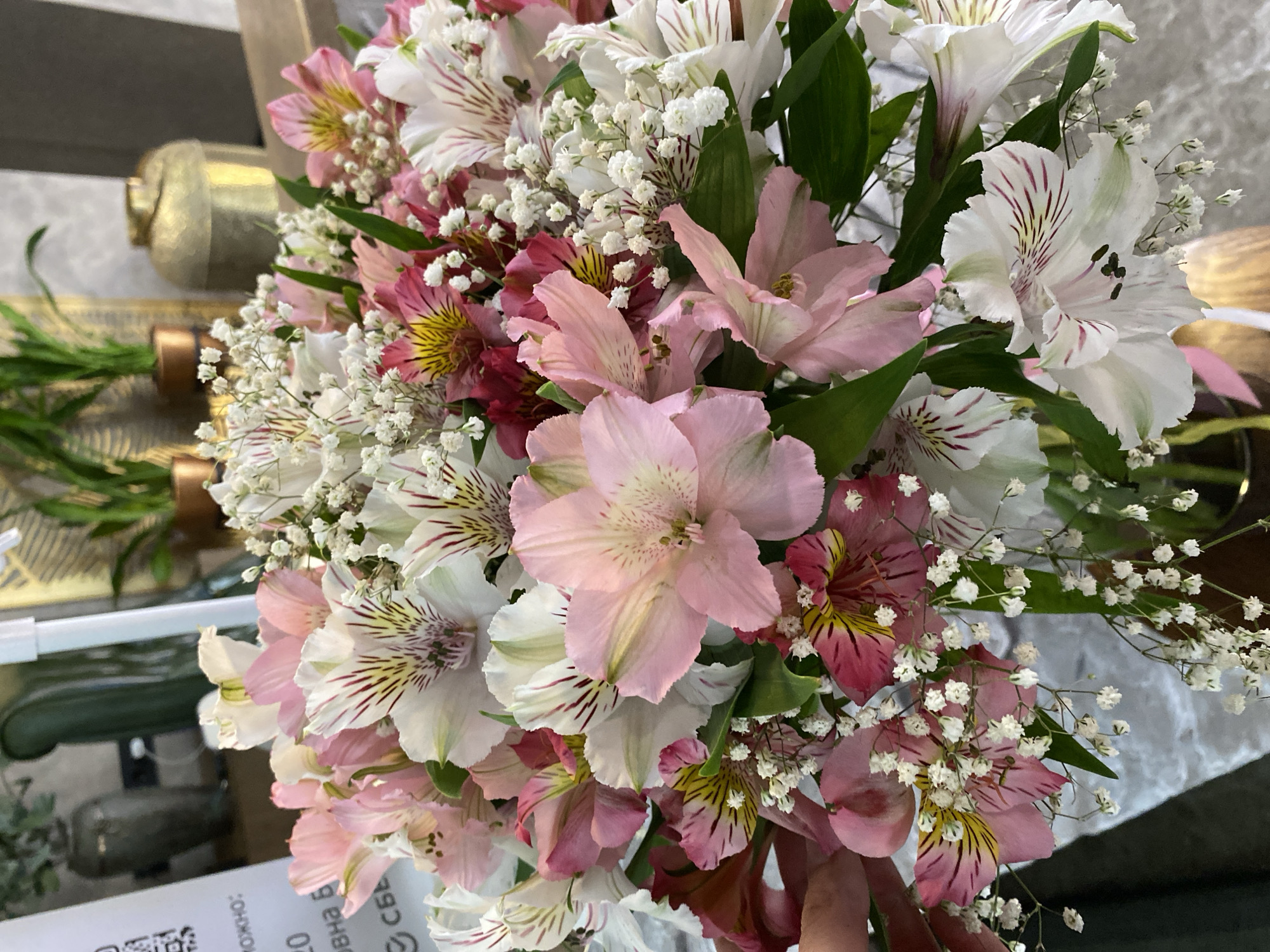 A close-up shot shows a bouquet of flowers, primarily composed of pink and white Alstroemeria and baby's breath. The flowers are in full bloom, with some blossoms displaying dark red stripes and yellow accents. The background is blurry but suggests an indoor setting. The presence of a golden object and what appears to be a QR code on a surface further suggests this is a commercial setting, such as a flower shop or event venue. There are no people explicitly visible in the scene. Given the request for location information in Votkinsk, Russia, it could be assumed that this bouquet is being displayed or sold at a floral establishment in the city.