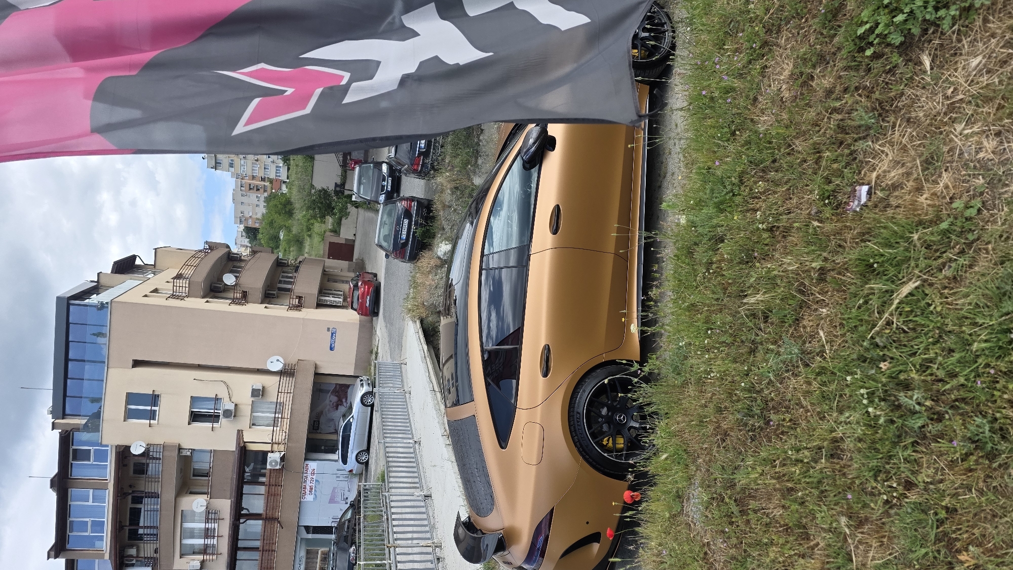 The image shows a matte gold car parked on the side of a street, with a portion of a black and pink flag visible in the upper left corner. The car is partially obscured by the flag and overgrown grass on the side of the road. In the background, there's a residential building with several balconies and satellite dishes. A street sign is visible on the building, marked "Serdika 18." Several other cars are parked along the street behind the featured car. The location is Blagoevgrad, Bulgaria. The environment appears to be residential with typical street-side parking. No people are visible in the image.