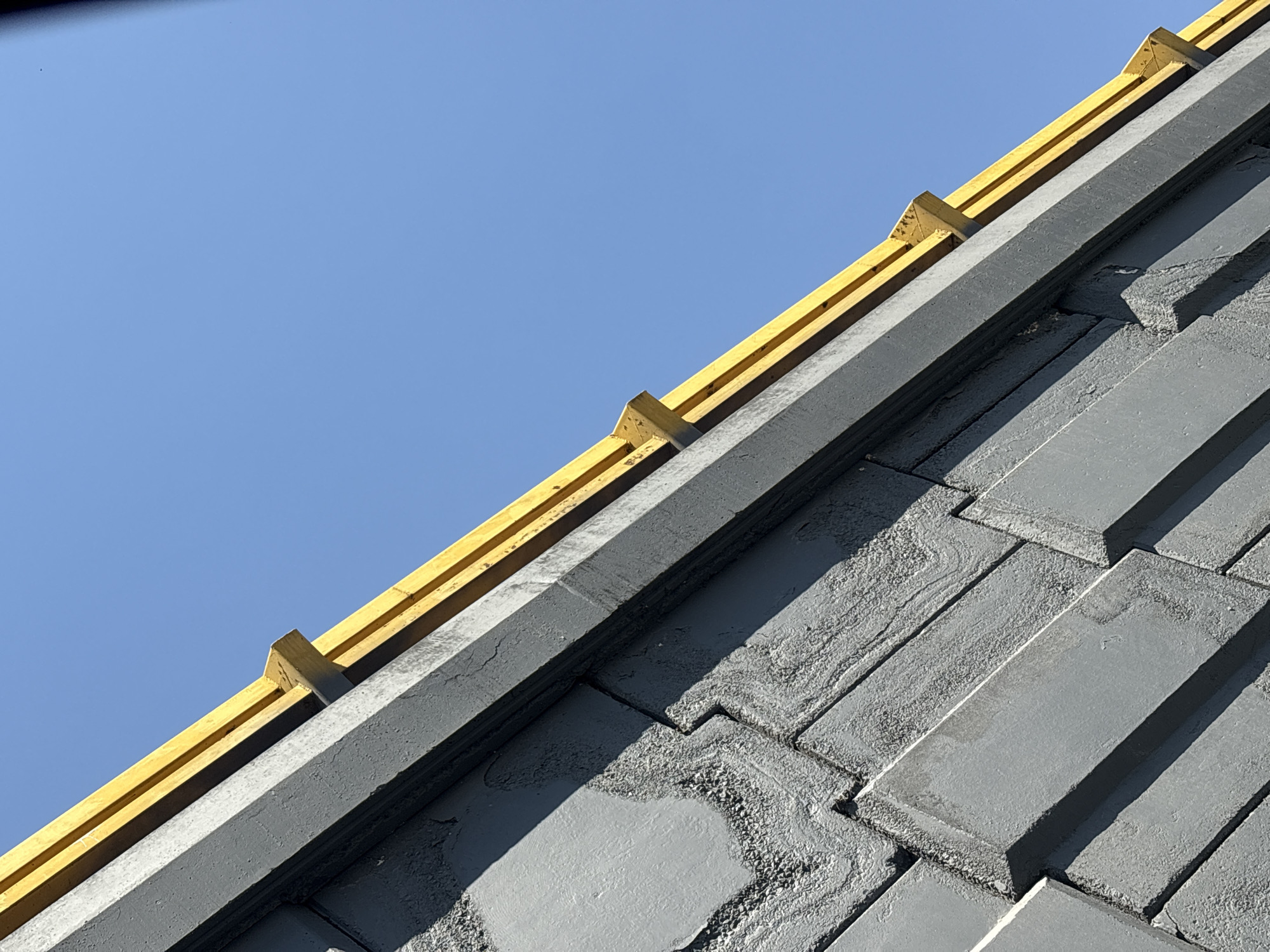 The image depicts a detailed view of a building's upper facade and roofline, situated in Puebla, Mexico. The lower portion of the frame features a textured, multi-toned gray wall or facade, characterized by stacked, rectangular, and irregularly shaped blocks. Some sections of these blocks display a wavy, almost fluid texture within the gray material. A distinct, smooth, dark gray architectural band or cornice runs diagonally from the lower left to the upper right, separating the textured wall from the elements above. Positioned atop this dark gray band are bright yellow architectural components: at least two parallel horizontal yellow strips and multiple smaller, wedge-shaped yellow supports projecting downward from the upper yellow strip. The background consists of a clear, unblemished light blue sky. Direct sunlight illuminates the scene, casting discernible shadows from the yellow elements onto the dark gray band and emphasizing the textures of the gray facade. No individuals, animals, or actions are present.