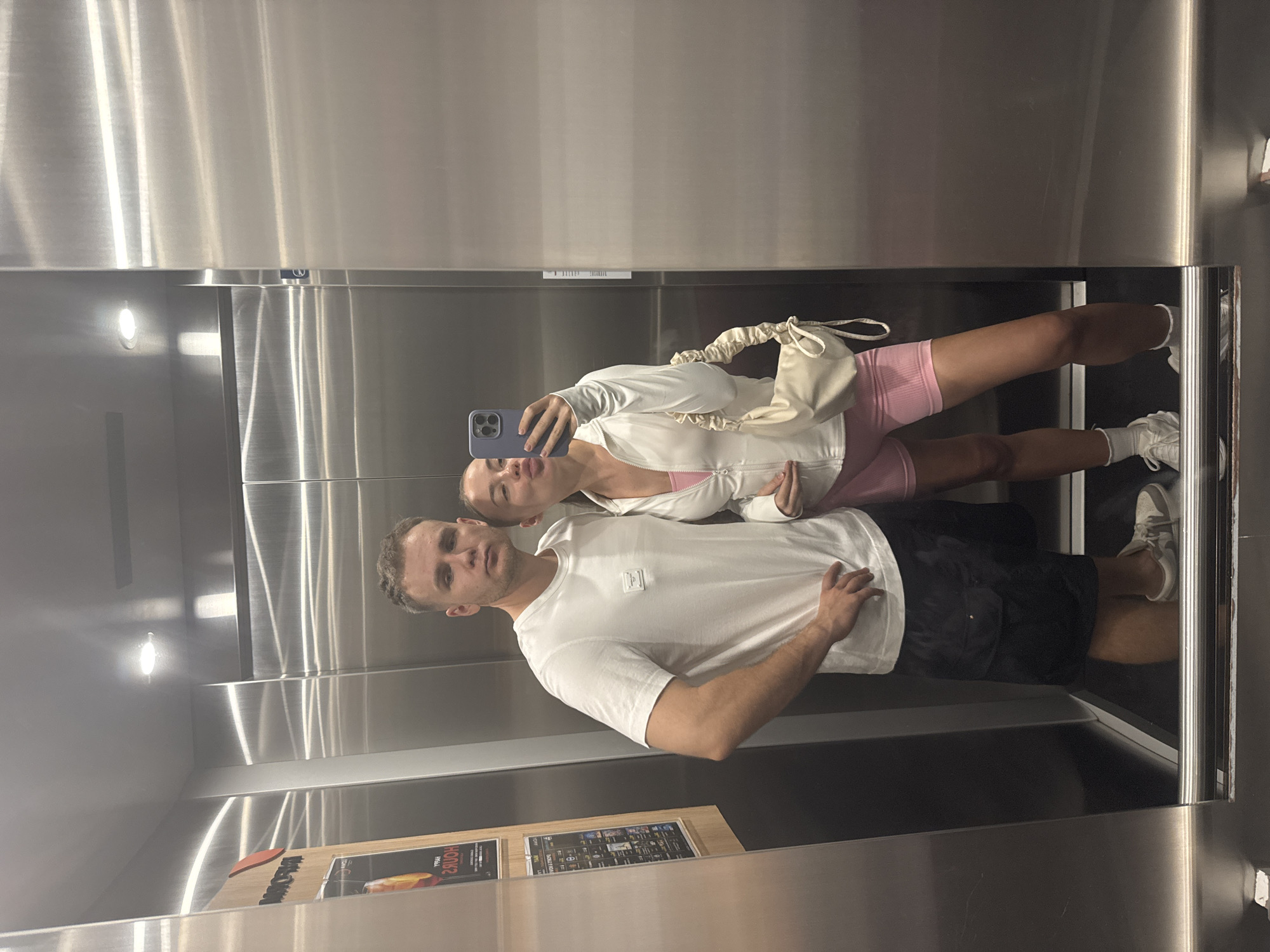A man and a woman are taking a selfie in an elevator. The man is wearing a white T-shirt and black shorts. The woman is wearing a white jacket and pink shorts, holding a blue smartphone, with a beige bag. The elevator has stainless steel walls and lighting. There is an advertisement on the wall near the floor. The scene appears to be located in a building in Choeng Thale, Thailand.