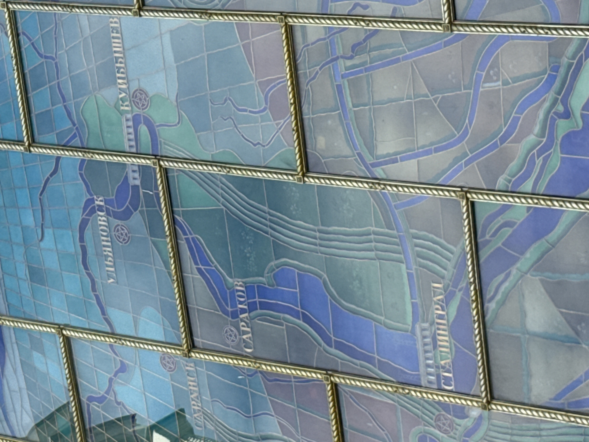 The image is a close-up of a large stained-glass mosaic depicting a map of Soviet-era cities along a river. The mosaic is divided into square panels framed by decorative metal trim. Several city names are visible in stylized lettering: "УЛЬЯНОВСК" (Ulyanovsk), "КУЙБЫШЕВ" (Kuybyshev), "САРАНСК" (Saransk), "САРАТОВ" (Saratov), and "СТАЛИНГРАД" (Stalingrad). The cities are marked with star symbols. The mosaic uses various shades of blue and green glass tiles to represent water and land. The overall impression is of a stylized, artistic representation of geography. The specific location of this mosaic in Moscow is not provided in the image itself.
