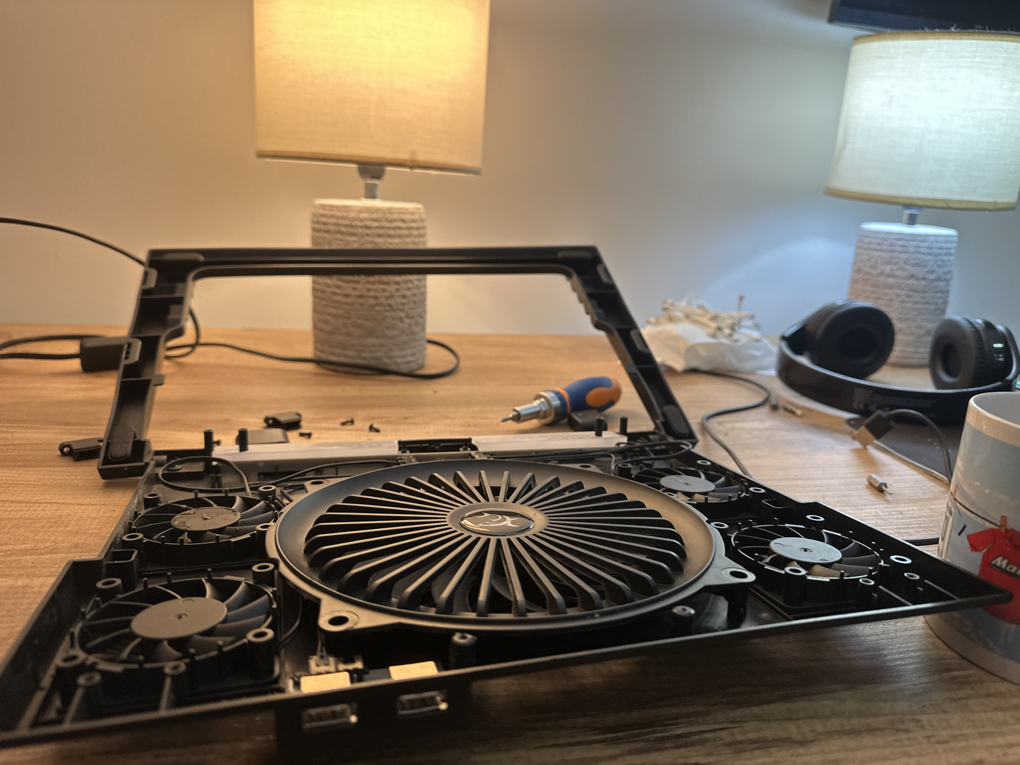 The image shows an open, disassembled laptop cooling pad on a wooden surface. The pad is primarily black plastic with multiple fans visible, including one large central fan with a silver logo. A blue and orange screwdriver sits nearby. Scattered around the cooling pad are small screws and electronic components. In the background, two lamps with neutral-colored shades sit on the same surface. A pair of headphones and a mug are also present on the right side of the frame. There is no information in the picture about the location, so it is impossible to know if it's in Budva, Montenegro.