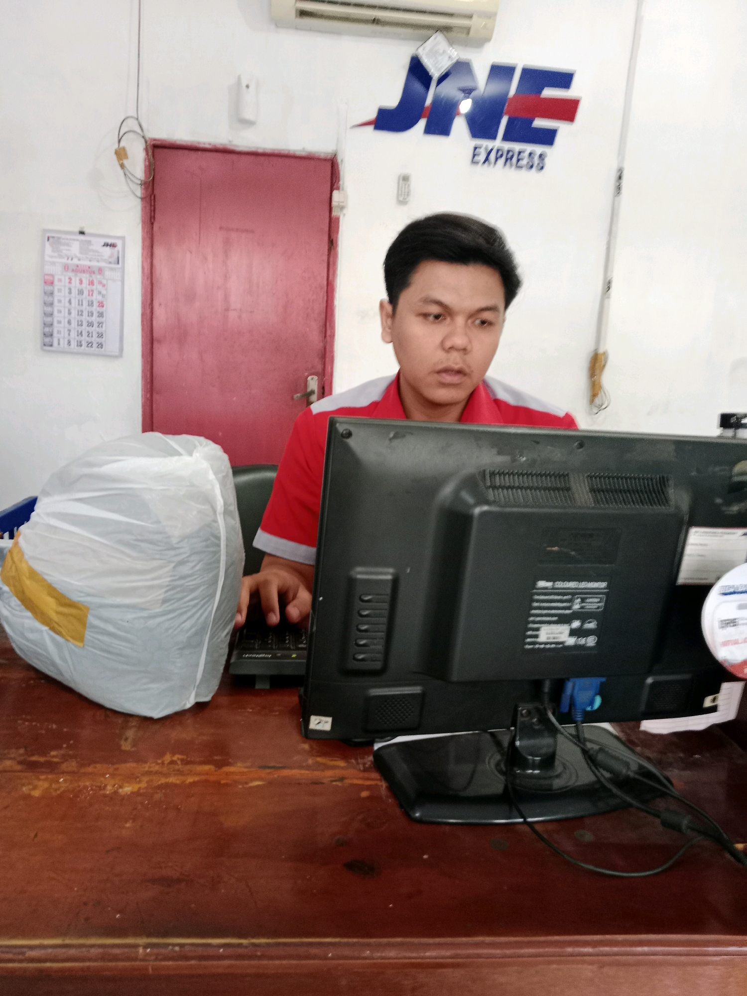 A young man, appearing to be in his early twenties, is seated at a wooden desk in what seems to be a JNE Express office in Probolinggo, Indonesia. He is wearing a red polo shirt with grey accents. His focus is directed towards a computer monitor in front of him, and his hands are on a black keyboard, suggesting he is working. To his left, there is a large, white plastic-wrapped object, possibly a package or a piece of equipment, sealed with yellow tape. Behind him, a red door is visible, along with a white calendar hanging on the wall to the left of the door, displaying dates. The JNE Express logo is prominently displayed on the wall behind the man. The overall environment suggests an operational office space.