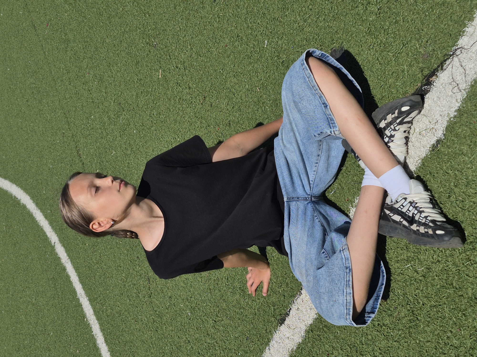 A young girl with light brown hair braided into a single plait sits cross-legged on a bright green artificial turf field. She is wearing a plain black t-shirt, loose-fitting light blue denim shorts that end above her knees, white socks, and black and white athletic shoes. Her eyes are closed, and she is looking down and to her right. A white painted line runs diagonally across the field, from the bottom left corner to the upper left of the frame. The background is out of focus, showing more of the green turf and a hint of a white line in the upper left. The lighting suggests it is a sunny day, with shadows visible on the turf. The image appears to have been taken outdoors in a sports facility, potentially a football or soccer field. This location is in the city of Chaikovsky, Russia.