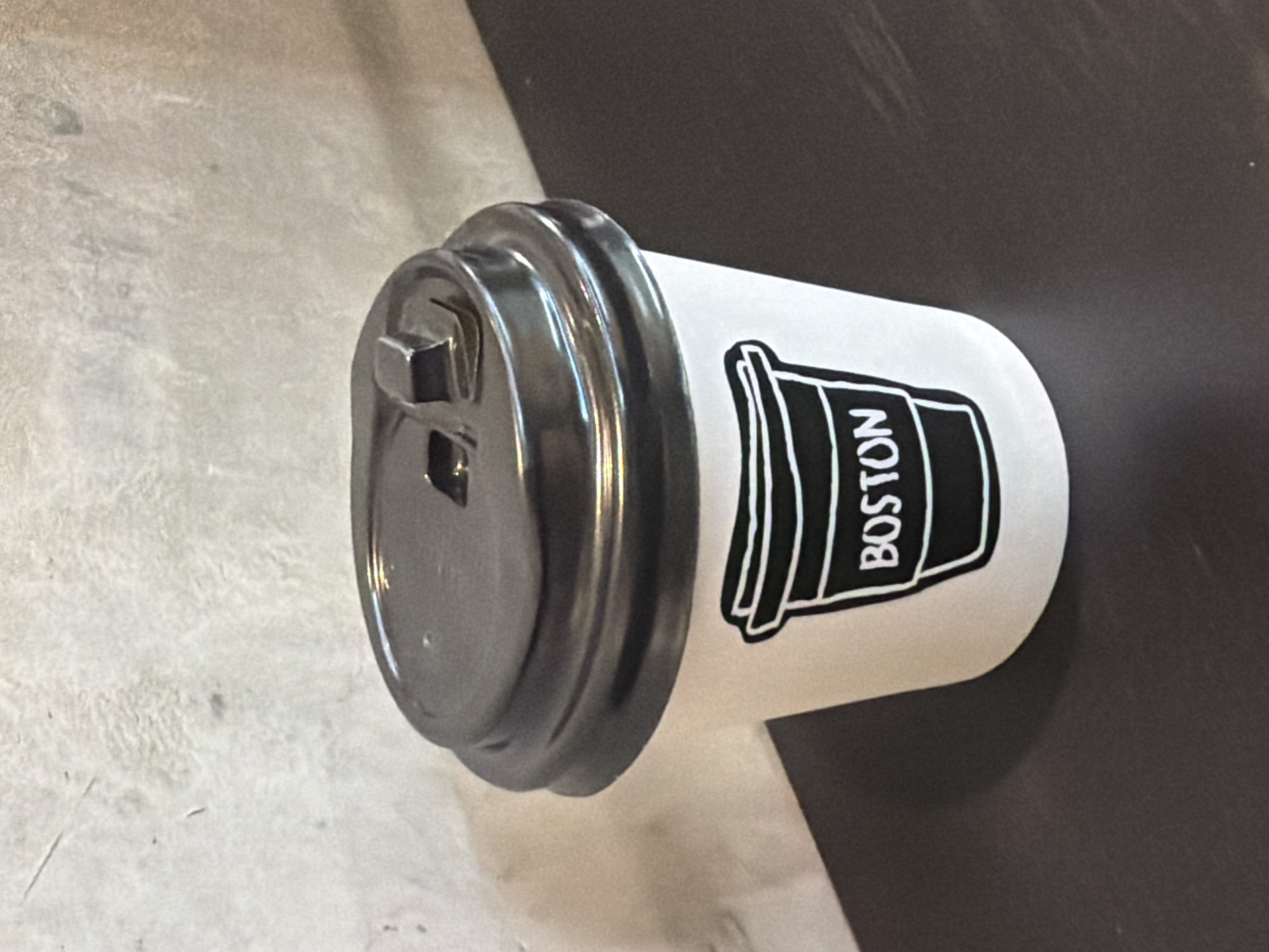 A white coffee cup with a black lid lies horizontally on a textured surface. The cup features a black outline graphic of a coffee cup on its side, inside which the word "BOSTON" is printed vertically in white capital letters. The black lid has a subtle sheen and includes a dark, square-shaped tab or opening mechanism.
The background surface is divided into two distinct regions. To the left of the cup, the surface is a light gray or off-white color, displaying a noticeable, somewhat rough texture, possibly indicative of concrete or a similar material. To the right of the cup, the surface is a uniform dark gray or black, appearing smooth and contrasting sharply with the light-colored cup.
There are no people or activities depicted in the image; the focus is entirely on the static object. The setting appears to be indoors, with no specific contextual details other than the text "BOSTON" on the cup, which suggests a connection to the city. The lighting is diffused, and the time of day is not discernible.