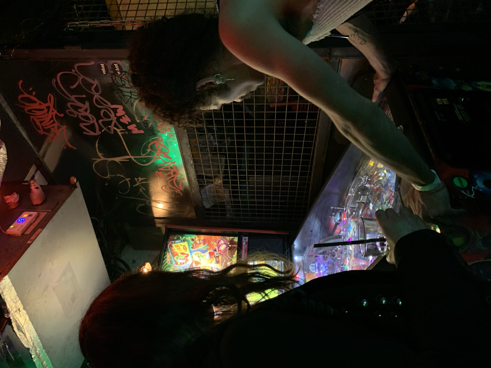 Two people play a pinball machine. One person with dark curly hair wears a white shirt and bracelet. The other person has long red hair and wears a black shirt. The pinball machine is brightly lit. The location is in Tel-Aviv, Israel. Graffiti is visible on a surface above the machine. A metal grid and other objects are nearby.