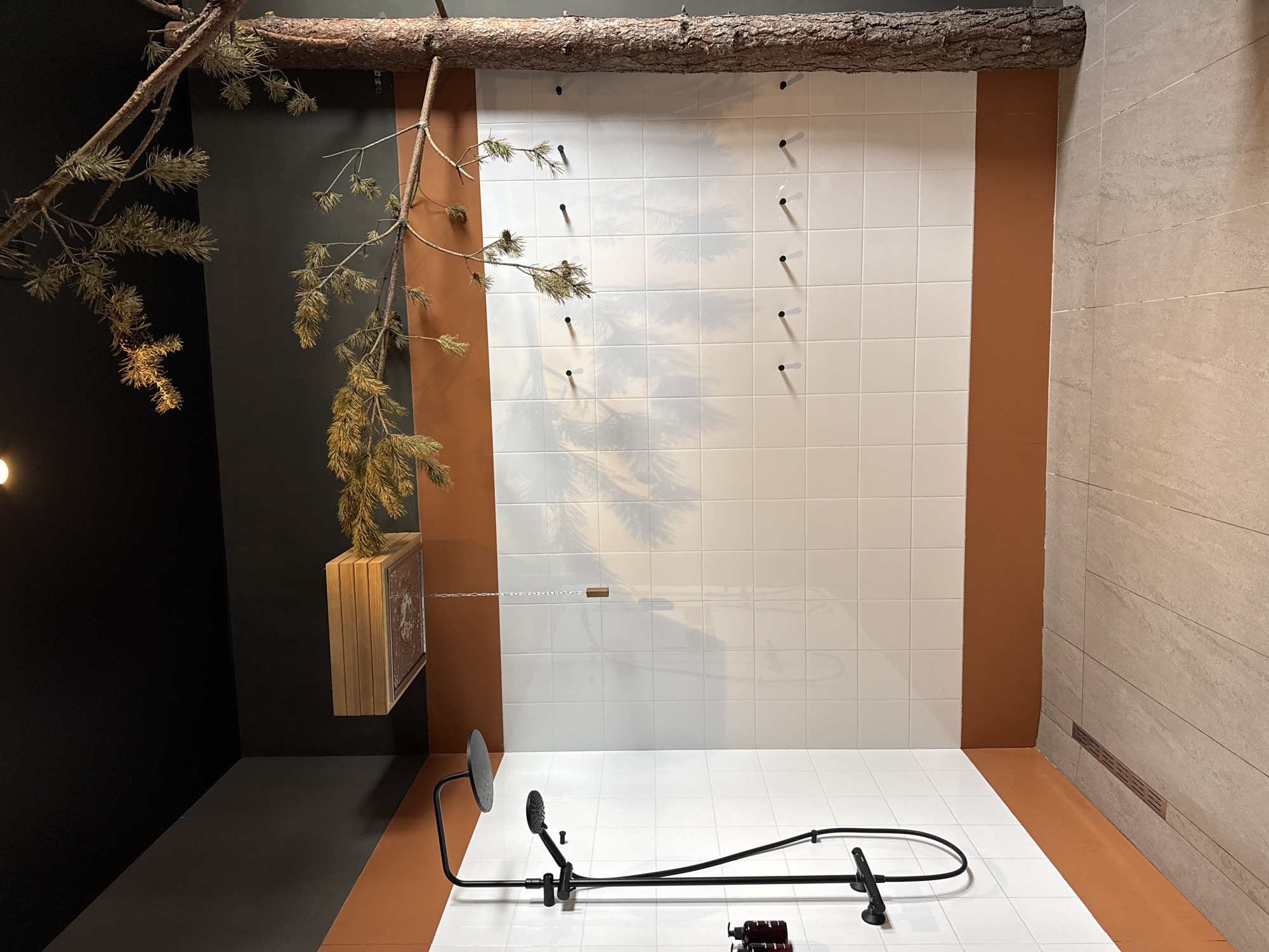 A showroom or bathroom display in Vyakhtelevo, Russia, features a shower fixture in a faux-natural setting. The walls are covered with white square tiles framed by brown borders. A weathered tree branch is mounted horizontally across the top, with smaller, dried branches hanging down. A small wooden box with decorative etchings is suspended from one of the branches. A black shower system with a large circular head and a smaller hand-held showerhead is installed on the tiled floor. A drain is visible at the edge where the tile meets the stone-like wall on the right. The floor is tiled in the same white square tiles as the wall. Small black pegs are attached to the wall, arranged vertically. The left side of the scene is dark, creating contrast with the white tiles. A shadow of the branches is projected onto the tiled wall. Brown bottles are visible near the base of the shower fixture.