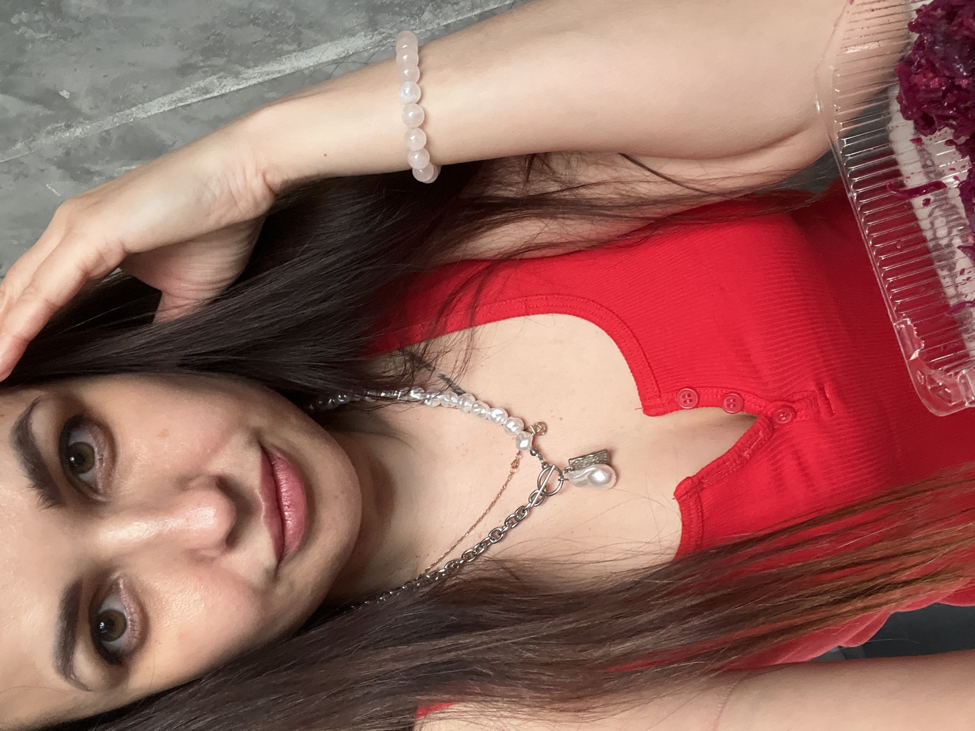 A woman is depicted in a close-up, upward-angled shot, possibly a selfie. She has long, dark brown hair, brown eyes, and dark eyebrows, with subtle makeup including light pink lipstick. She is wearing a bright red, ribbed top with a scoop neckline and a placket featuring three small red buttons. Around her neck are two layered necklaces: one is a silver-toned chain with a toggle clasp and a large teardrop-shaped pearl pendant, and the other is made of irregular white pearls, layered with a thin, rose gold-colored chain that has a tiny charm. On her right wrist, a bracelet made of light pink, spherical beaded stones is visible. Her right hand is partially visible near her head, holding or resting on her hair. In the upper right corner, a clear plastic container holds a dark reddish-purple, textured food item, likely a dessert. The background is a textured, mottled grey surface, suggesting an indoor setting. The woman has a soft, pleasant expression, looking directly at the camera. There is no visible text.