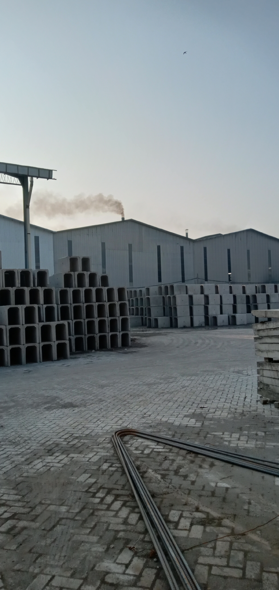 The image shows a concrete manufacturing facility in Sumput, Indonesia. Stacks of concrete culverts and flat slabs are visible in the foreground and midground, along with a coil of rebar resting on a brick-paved yard. In the background, multiple prefabricated metal buildings with smoke emanating from one chimney are present. An overhead crane structure is visible on the left, and a single bird is in the sky.