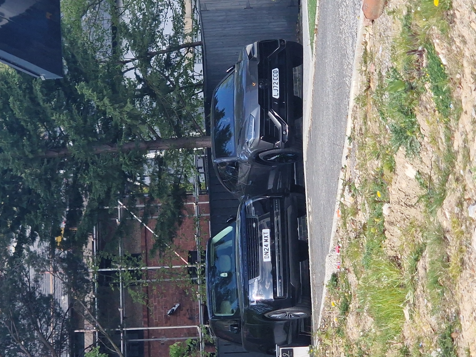 The image captures two luxury black SUVs parked outdoors in Witton Gilbert, United Kingdom, under bright daylight, suggesting a clear and sunny day. The scene shows the vehicles positioned on what appears to be a paved area or driveway, adjacent to a sidewalk and a strip of unkempt ground.
The vehicle on the right is a black Lamborghini Urus, clearly displaying the UK registration plate "LJ72 CCO". Its aggressive front grille and styling are prominent. The vehicle on the left is a black Range Rover, identifiable by the "RANGE ROVER" text across its bonnet, and bearing the UK registration plate "DN24 MXT".
In the background, dense green foliage from trees dominates, partially obscuring a brick building. Scaffolding is visible against the brickwork of the building, suggesting ongoing construction or renovation. A dark wooden fence or building facade is situated directly behind the Lamborghini.
The foreground and right side of the image show a paved sidewalk or path bordering a strip of dry earth mixed with green grass and small weeds, some bearing tiny yellow flowers. No people are visible, and the primary activity captured is the stationary presence of the two vehicles.
Visible text includes the two distinct UK license plates: "LJ72 CCO" on the Lamborghini Urus and "DN24 MXT" on the Range Rover. The "RANGE ROVER" branding is also clearly seen on the second vehicle. A partial, blurry number "2" can be faintly seen in the bottom right corner of the original image. The overall setting appears to be a residential or semi-urban area.