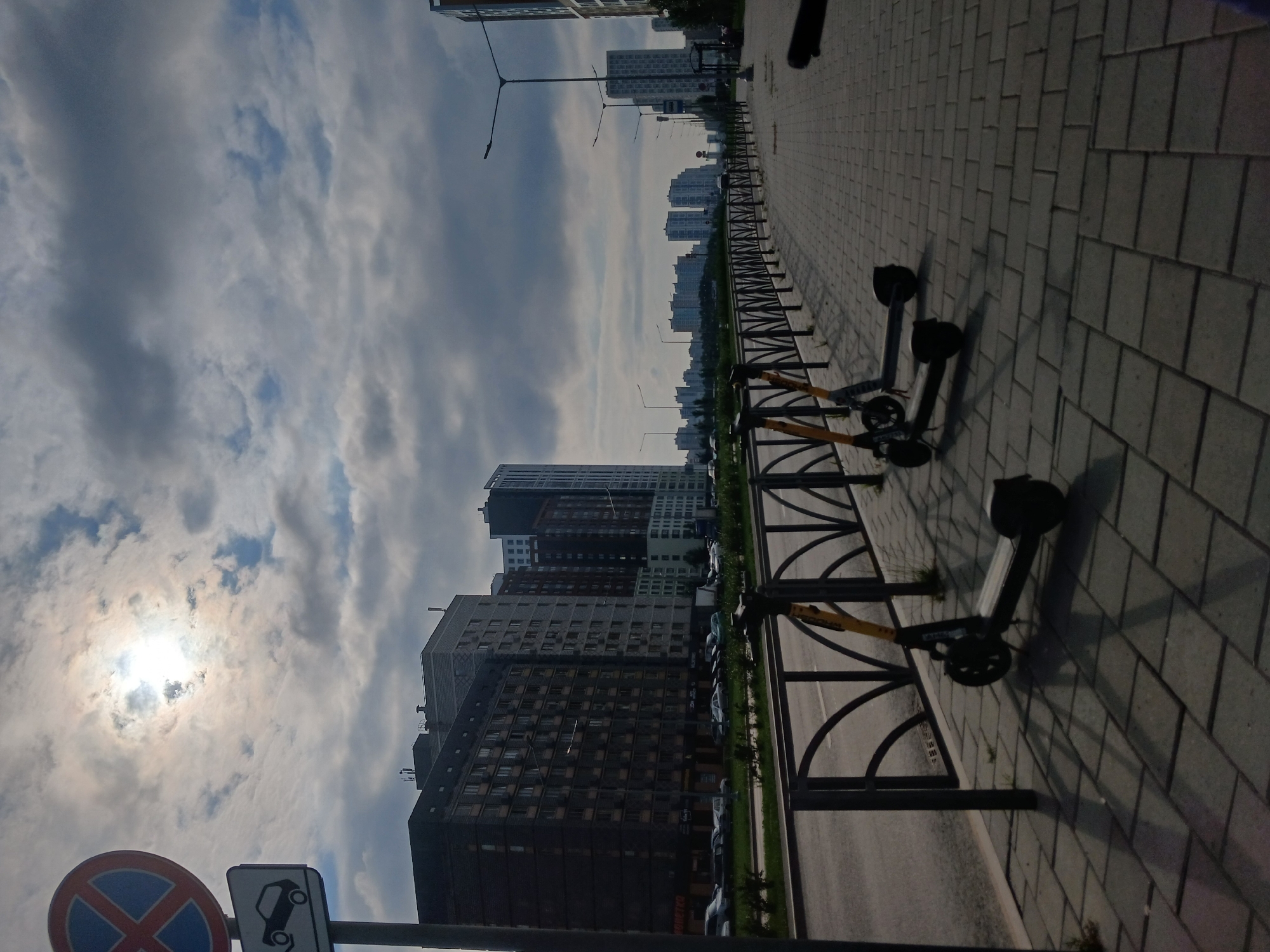The image depicts a street scene in Yekaterinburg, Russia, on a cloudy day. In the foreground, three electric scooters are parked along a paved sidewalk lined with a black metal fence. The scooters are black with yellow accents. Behind the fence is a road with a few cars visible and a grassy strip. Several tall apartment buildings form the backdrop, with a mix of dark and lighter colored facades. The sun is partially obscured by clouds, casting dappled light and shadows. A traffic sign with a red circle and a diagonal bar, indicating no stopping, is visible on the left side of the image, along with a secondary sign showing a car icon with a red diagonal bar. The sky is filled with dramatic, heavy clouds.