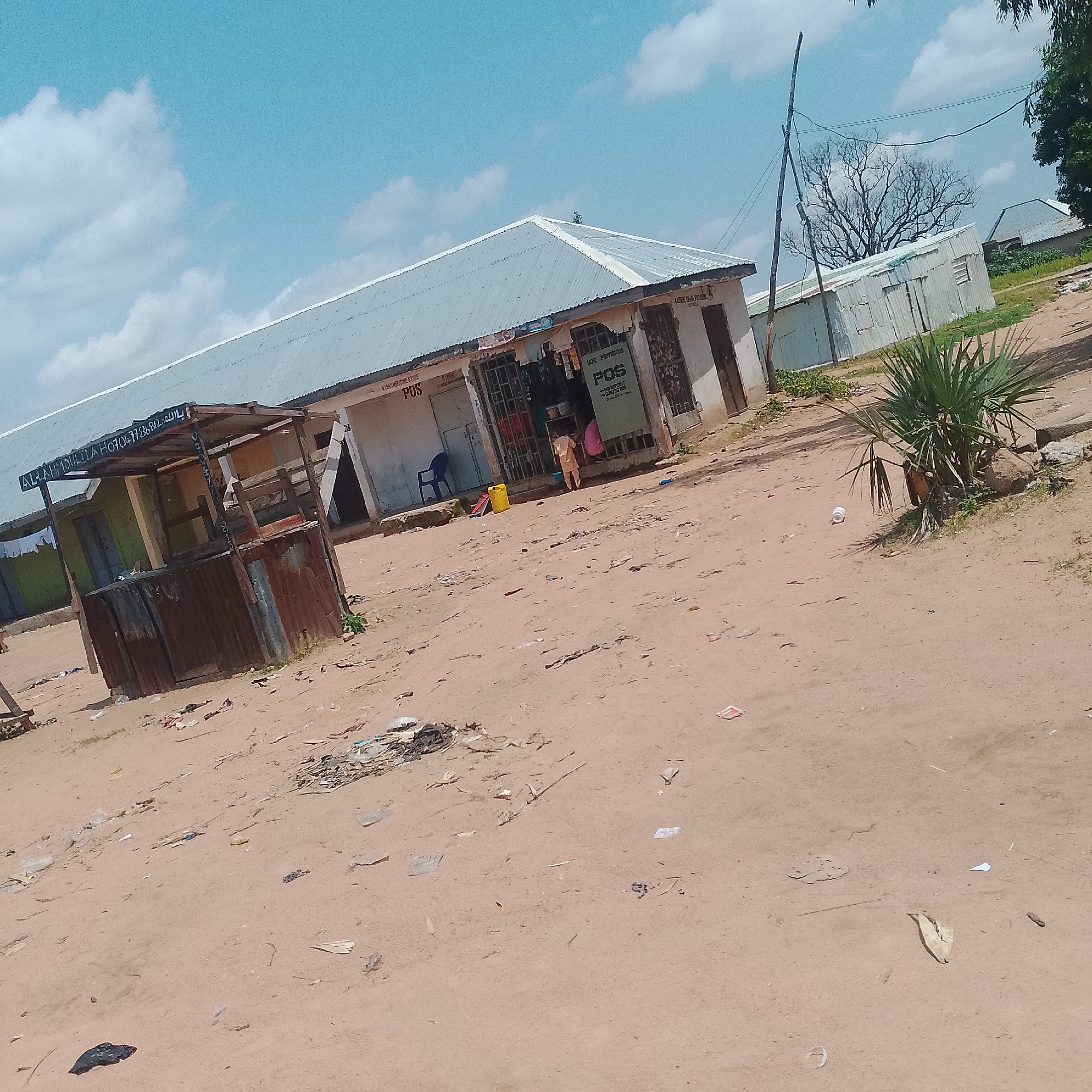 The image depicts a dusty street scene in Jalingo, Nigeria, with a cluster of shops and makeshift stalls. In the foreground, a rustic, open-sided stall constructed with wood and corrugated metal stands, appearing weathered and somewhat disused. White lettering on a vertical support post reads "ALLAHMDULLILA" and a phone number.
Further back, a longer, more permanent structure houses several businesses. A sign above one section reads "KASAD MEDICINE STORE POS." Another section displays "CORE PROVISIONS POS" with some handwritten text below. Through an open, barred entrance, shelves of goods are visible. A child in a light-colored outfit stands near this entrance, facing away from the camera. A yellow jerrycan is placed outside the store. A blue plastic chair is visible to the left of the entrance.
The ground is predominantly sandy and dirt, littered with various pieces of debris and trash, including plastic bags, paper scraps, and other unidentifiable items. A single white plastic cup is visible on the ground in the middle distance, near a bushy plant with spiky leaves. In the background, other corrugated metal structures and a taller building with a similar roofline are present. A wooden utility pole with wires stretches across the upper portion of the scene. The sky is bright blue with scattered white clouds. The overall impression is of a modest, somewhat informal commercial area in a developing urban or semi-urban environment.