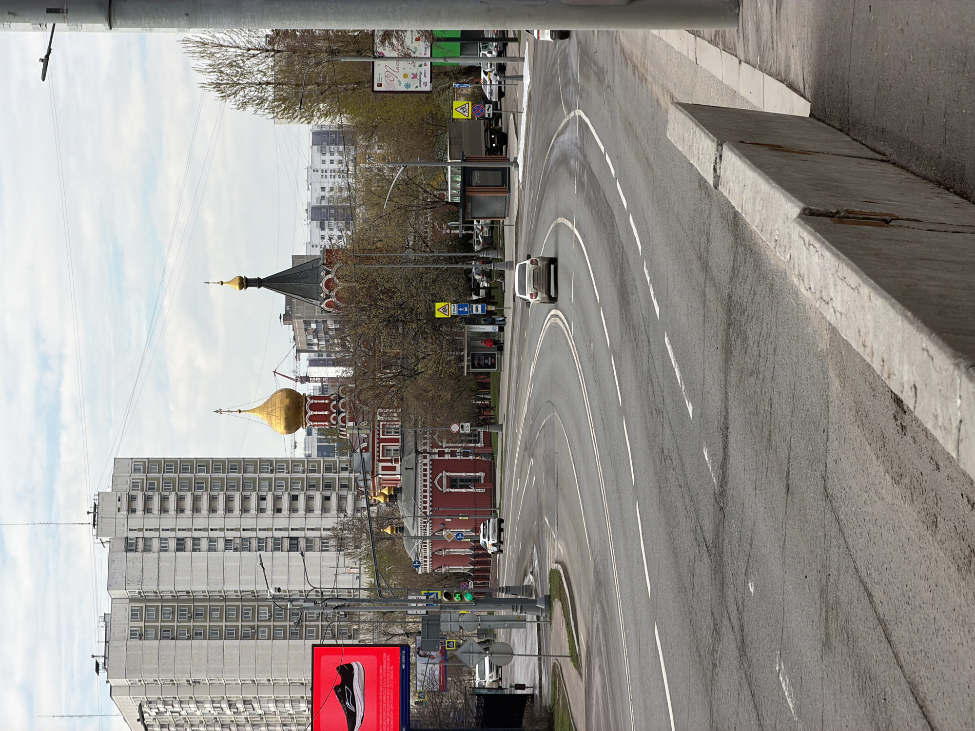 The image shows a street view in Moscow, Russia. A road curves to the left, marked by white lane lines, with a white car driving on it. A concrete barrier separates the road from the sidewalk. In the background, a Russian Orthodox church with gold domes stands out amidst city buildings. A large advertising billboard displays a shoe ad, and traffic lights are visible at an intersection. The sky is overcast with a few clouds. Trees with budding leaves are present along the street.