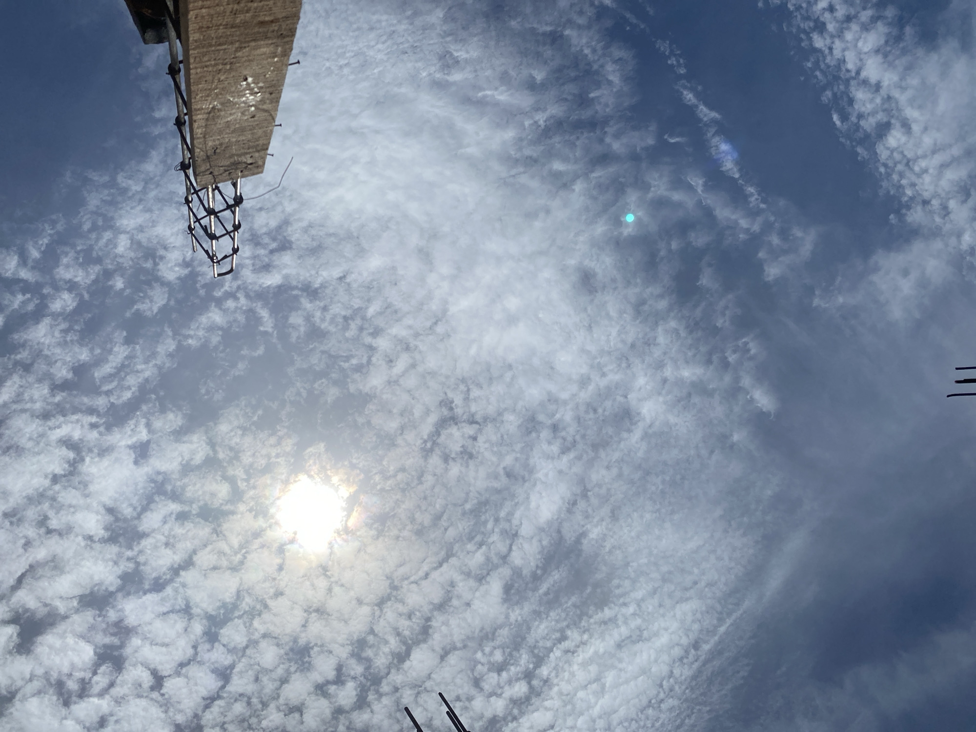 The image is an outdoor, low-angle shot looking up at a bright, partly cloudy sky. The sun is visible, casting a bright glare and creating a rainbow effect. There are scattered clouds, some appearing puffy and others wispy. In the upper right corner, a portion of a wooden structure with metal reinforcement is visible. On the left side of the image, two dark, vertical objects protrude into the frame. The overall impression is a construction site or an urban area with a clear sky. There are no people or animals in the image. The environment appears to be an open area, possibly under construction.