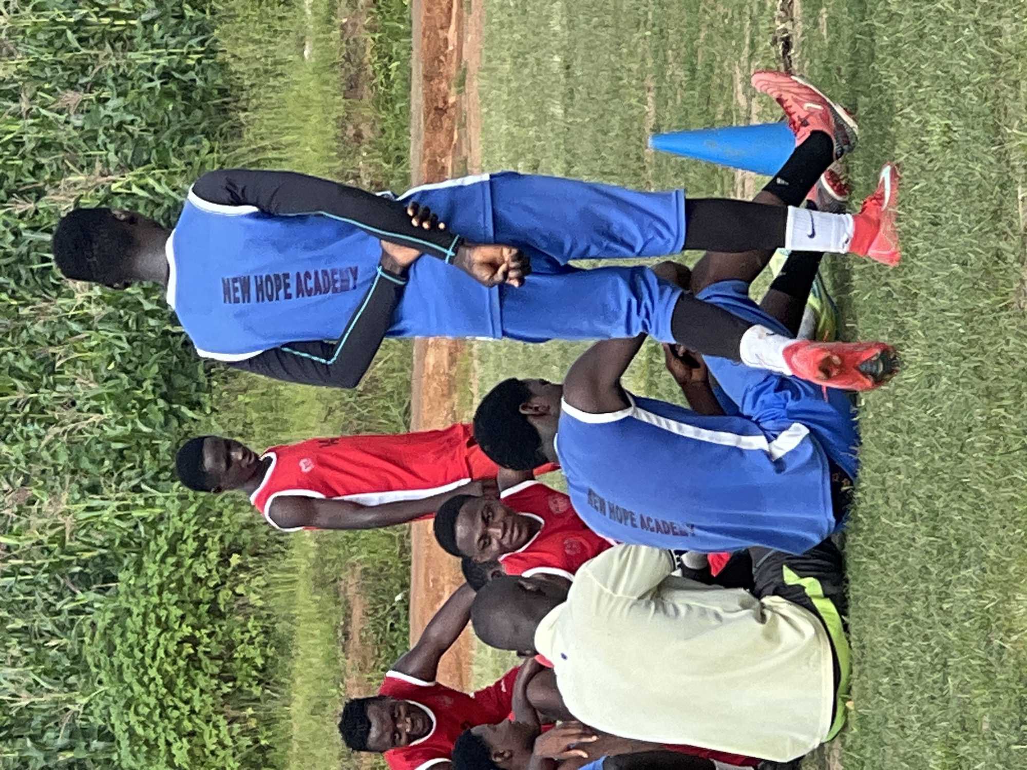 In an outdoor scene on what appears to be a grassy sports field in Jos, Nigeria, a group of young men, likely a sports team, are gathered. Some are seated on the ground, while others stand. The majority are wearing athletic attire, predominantly red and blue jerseys with the inscription "NEW HOPE ACADEMY" visible on the back of one blue jersey. The environment is characterized by lush green vegetation in the background, possibly corn stalks, and a reddish-brown dirt path. One individual in the foreground is wearing a light yellow shirt. A blue traffic cone is visible on the right side of the image. The lighting suggests it is daytime.