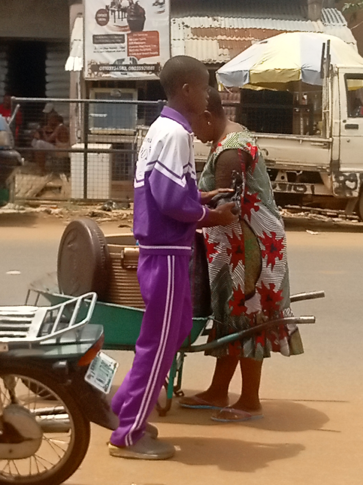 In Ogidi, Nigeria, two individuals are positioned on a street next to a motorized vehicle. One is a young person dressed in a purple tracksuit with white stripes, while the other is a woman wearing a dress with a pattern featuring red floral motifs. A wheelbarrow laden with goods is situated between them. The area contains buildings and vehicles; a truck with an umbrella-like covering and an advertisement board are visible in the backdrop.