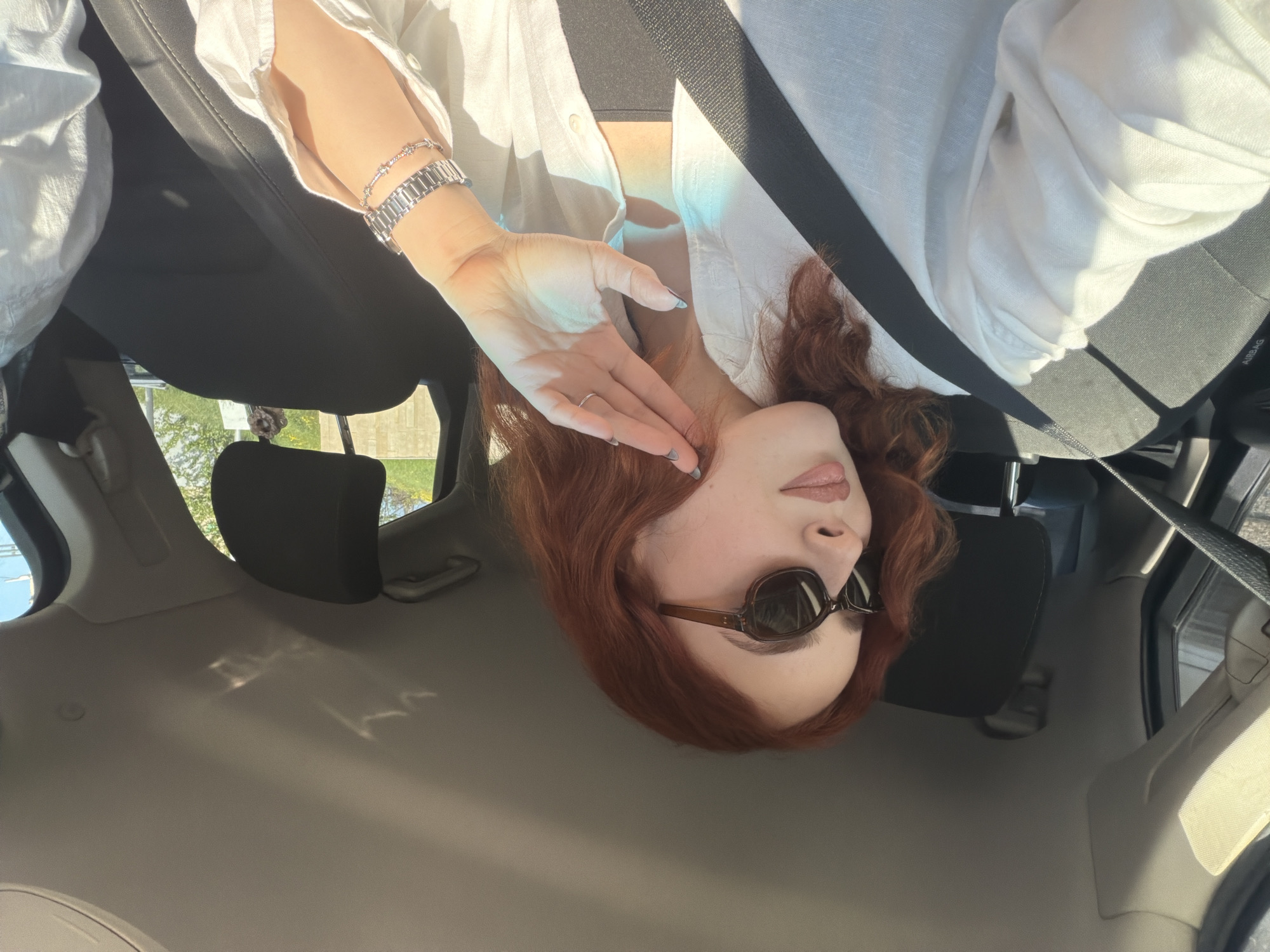 The image depicts a person sitting in the passenger seat of a car on a sunny day. The shot is taken from a slightly low angle, looking upwards towards the subject. The setting is inside a car in Tbilisi, Georgia, under bright daylight conditions.
The main subject is a person with reddish-brown, wavy hair, wearing dark, oval-shaped sunglasses and a light-colored, possibly white or off-white, linen button-up shirt. The shirt is unbuttoned to reveal a black top or bra underneath. One hand is gently placed near their face, with fingers touching the chin and cheek. This hand features a silver-toned ring on the ring finger and nails painted with a light blue polish. On the other wrist, a silver-toned watch with a link band is worn, accompanied by a thinner, braided silver bracelet.
Bright sunlight streams into the car, creating strong highlights on the white shirt and skin, and casting distinct shadows. The sunlight also produces an iridescent blue reflection or tint in certain areas, particularly on the shirt and the hand. The person is wearing a black seatbelt across their chest, which passes over the white shirt.
The car's interior is visible, including the dark headrest of the passenger seat, parts of the light-colored headliner, and a portion of the car's window showing glimpses of green foliage and a clear sky outside. On the car's interior, likely on the side of the seat or an adjacent panel, the text "AIRBAG" is faintly visible. The overall impression is one of casual comfort and relaxation in a sunlit environment.