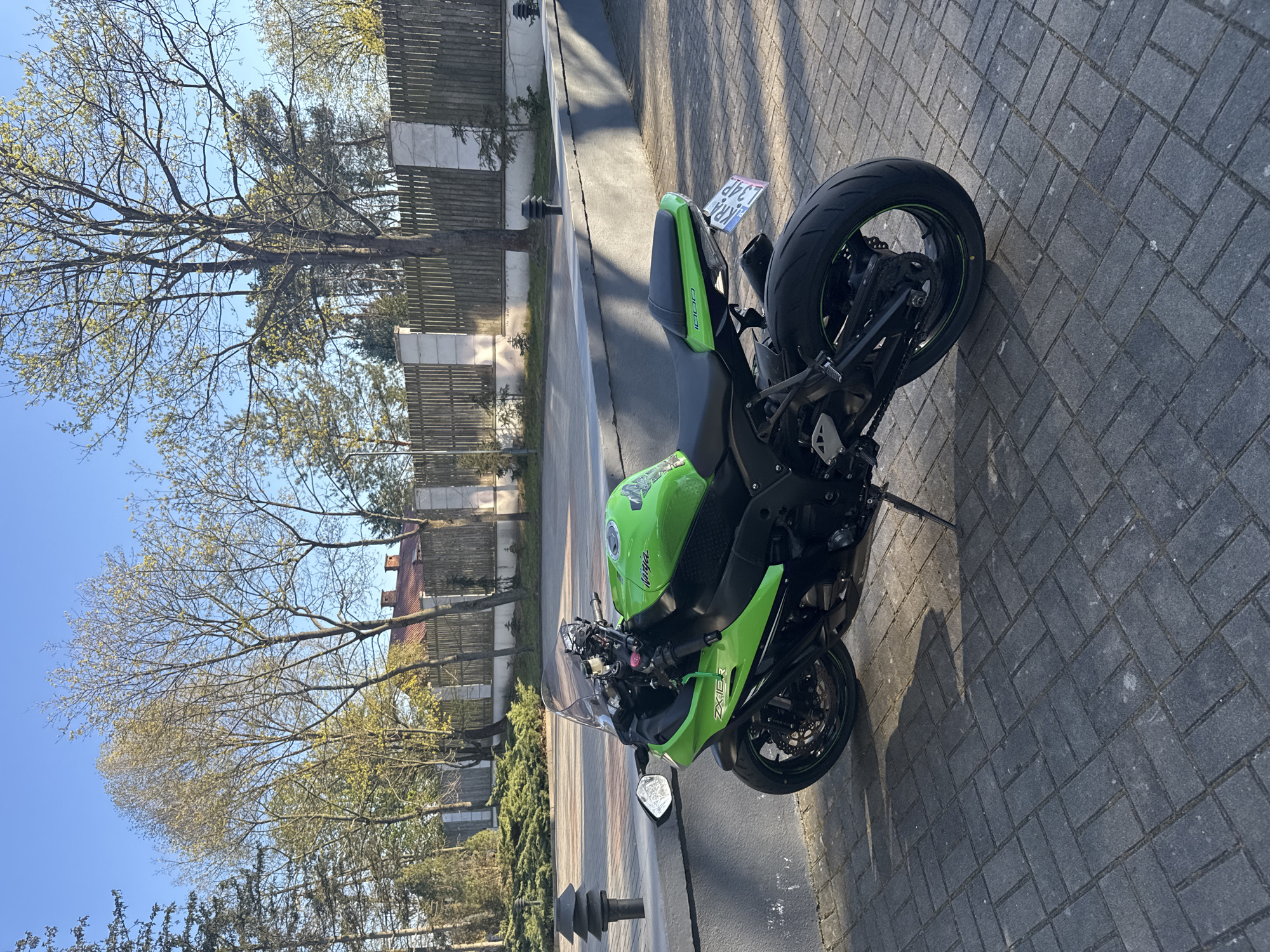 A Kawasaki motorcycle, predominantly green and black, is parked on a gray brick paved area alongside a street. The motorcycle is leaning on its side stand. Its license plate reads "KRA L34P." Behind the motorcycle is a low concrete wall with sporadic decorative black posts. A wooden fence runs parallel to the wall, with trees and shrubs growing behind it. Further back, a building with a reddish-brown roof is partially visible. The sky is blue. The location is Białobrzegi, Poland.