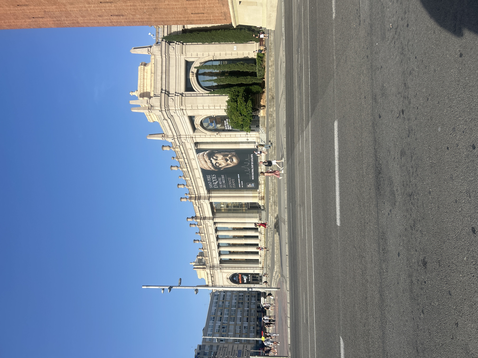 The image captures a wide shot of the National Art Museum of Catalonia (Museu Nacional d'Art de Catalunya) located on Montjuïc hill in Barcelona, Spain. The building, characterized by its neoclassical architecture, features a grand facade with numerous columns and arched windows. A large banner displaying a sculpture head and text is prominently displayed on the building's exterior, indicating an ongoing exhibition.
In front of the museum, a wide paved area serves as a pedestrian walkway and access point, with a street and a road with white lane markings in the foreground. Several individuals are seen walking on the plaza and stairs leading to the museum. The sky is clear and blue, suggesting a sunny day. To the right, a portion of a tall, red brick structure, likely part of the Montjuïc Palace, is visible. The scene is bathed in natural light, casting shadows on the ground.
